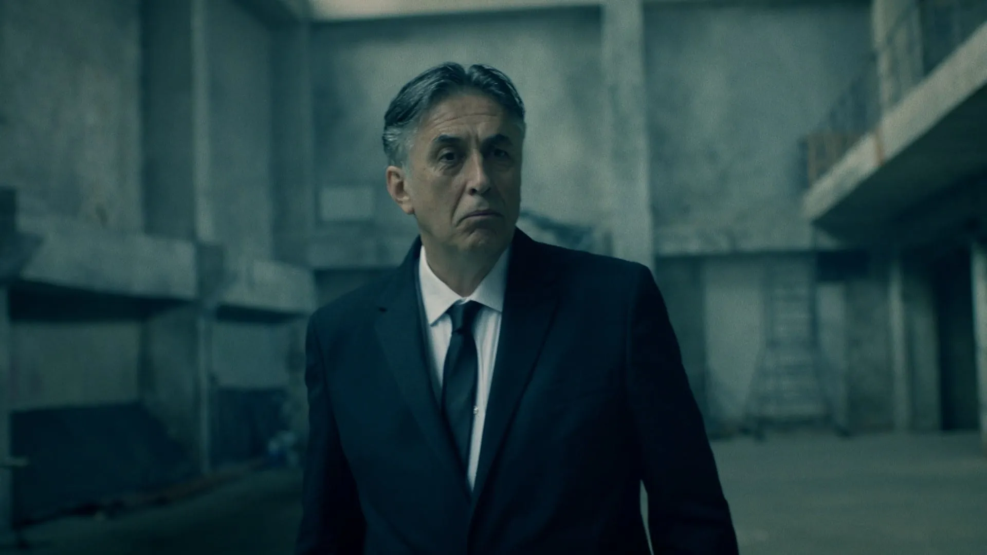 A grey-haired man in a dark suit walks through an empty concrete hall, staring straight ahead
