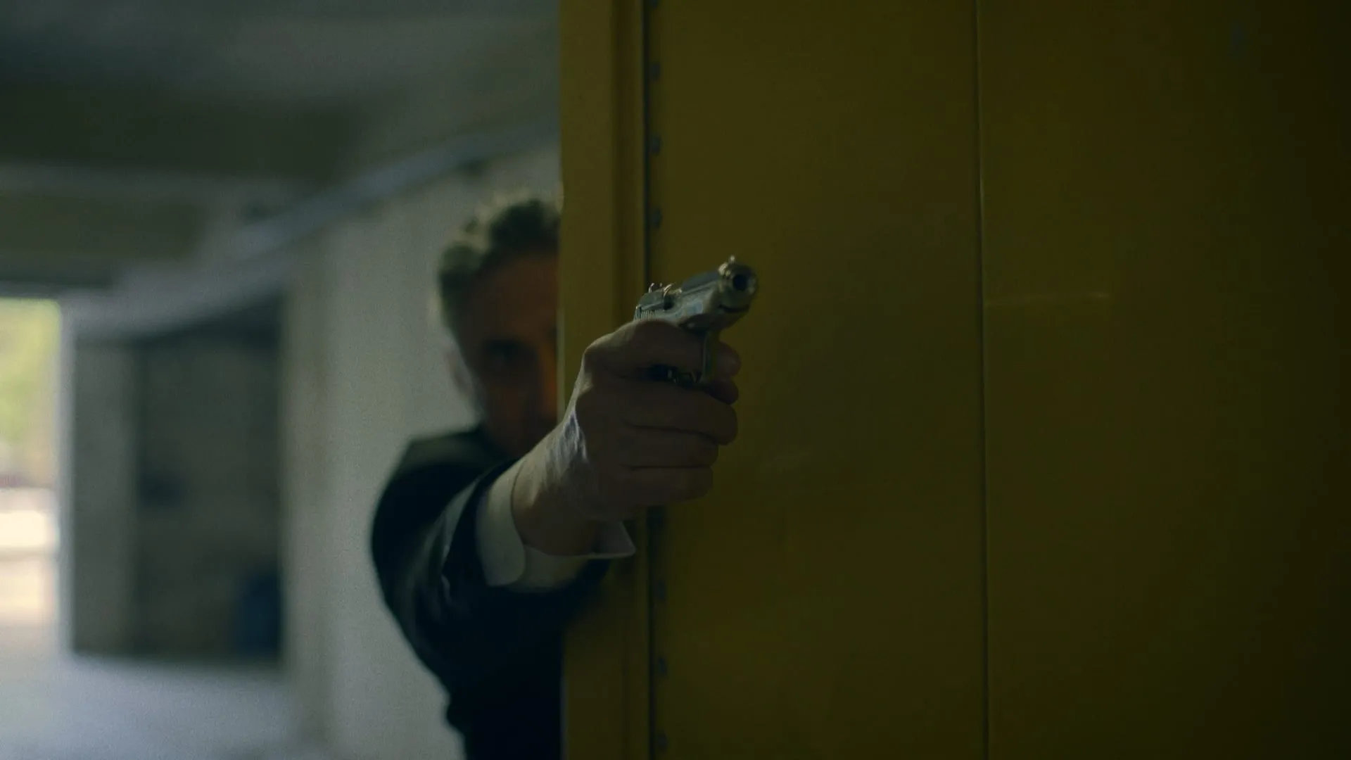 A man in a dark suit leans out from behind a yellow pillar, pistol aimed down the corridor