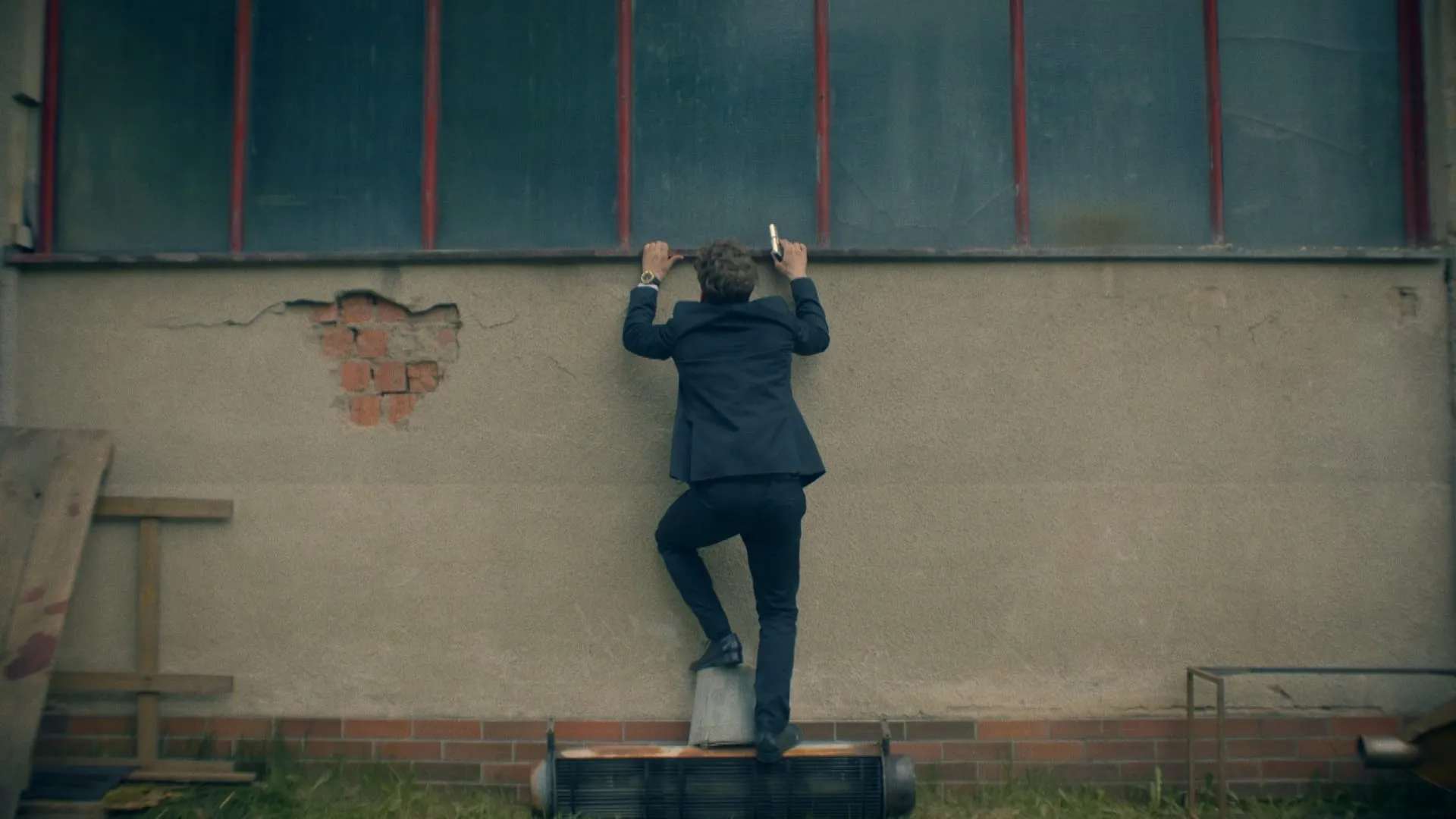 A man in a suit climbs a wall towards high windows, standing on a barrel with a phone in his hand