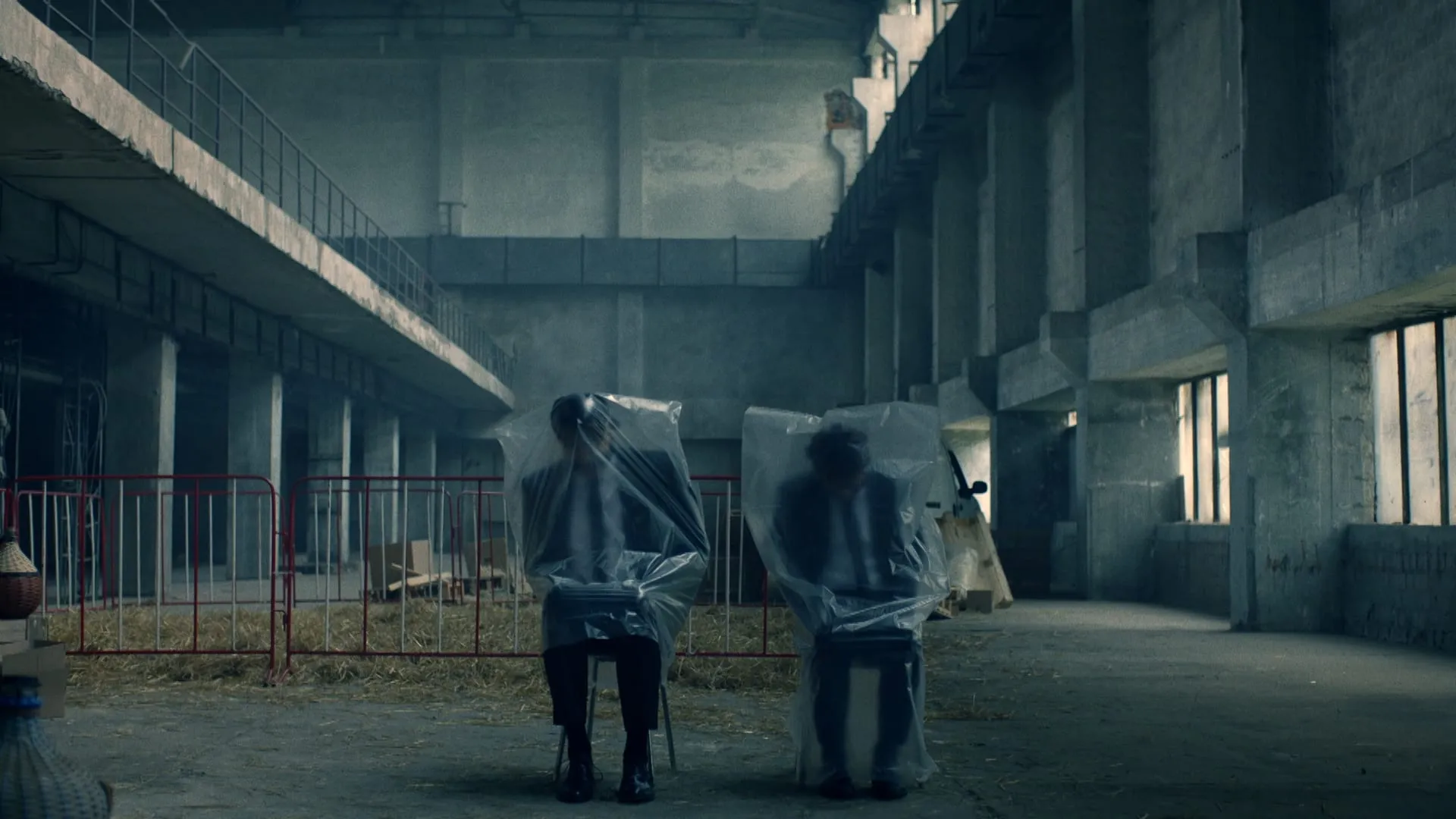 Two men in suits sit with plastic sheeting over their heads in an abandoned concrete hall