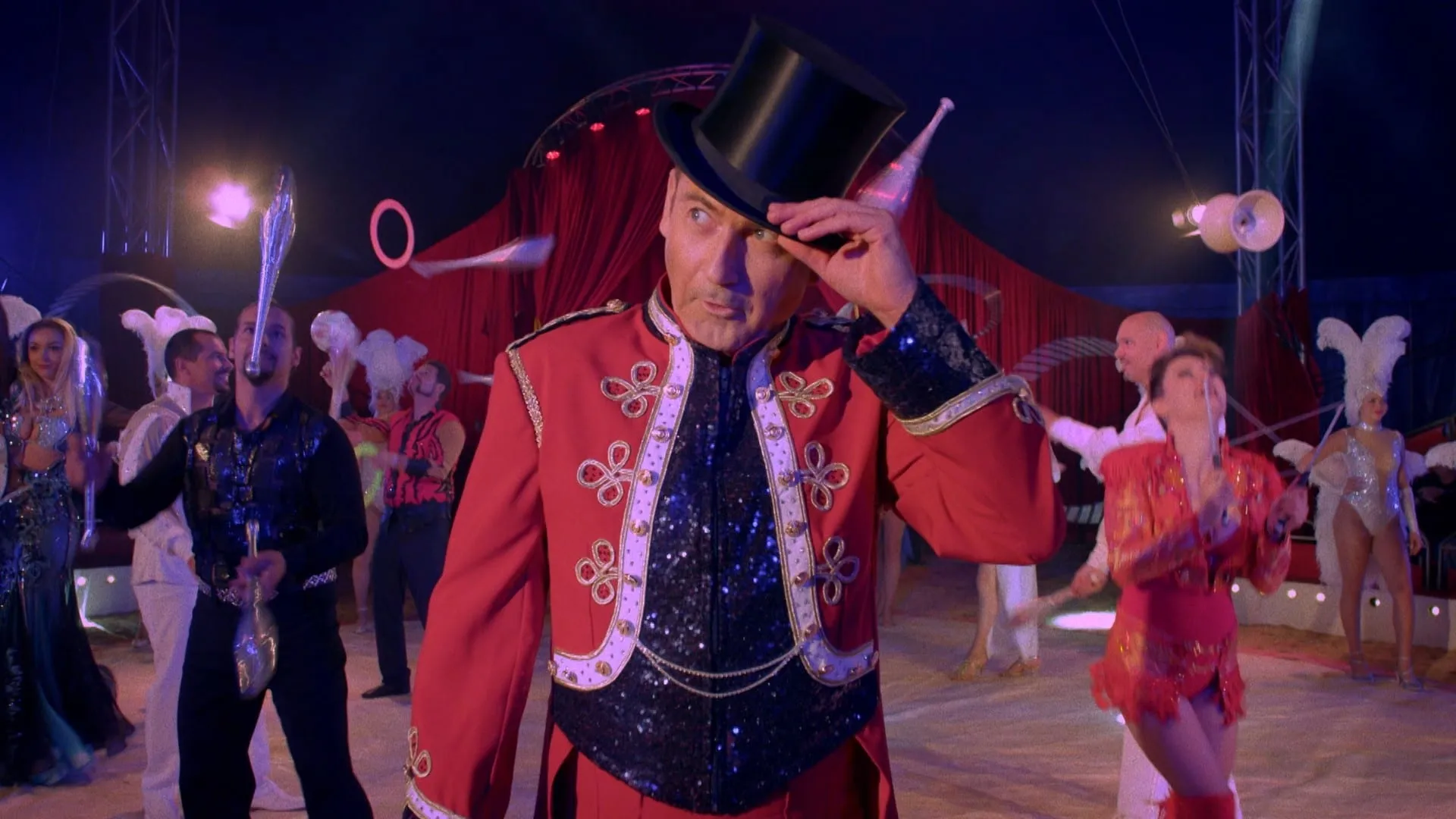 A ringmaster in a red braided tunic steadies his top hat in the ring as artists juggle clubs behind him