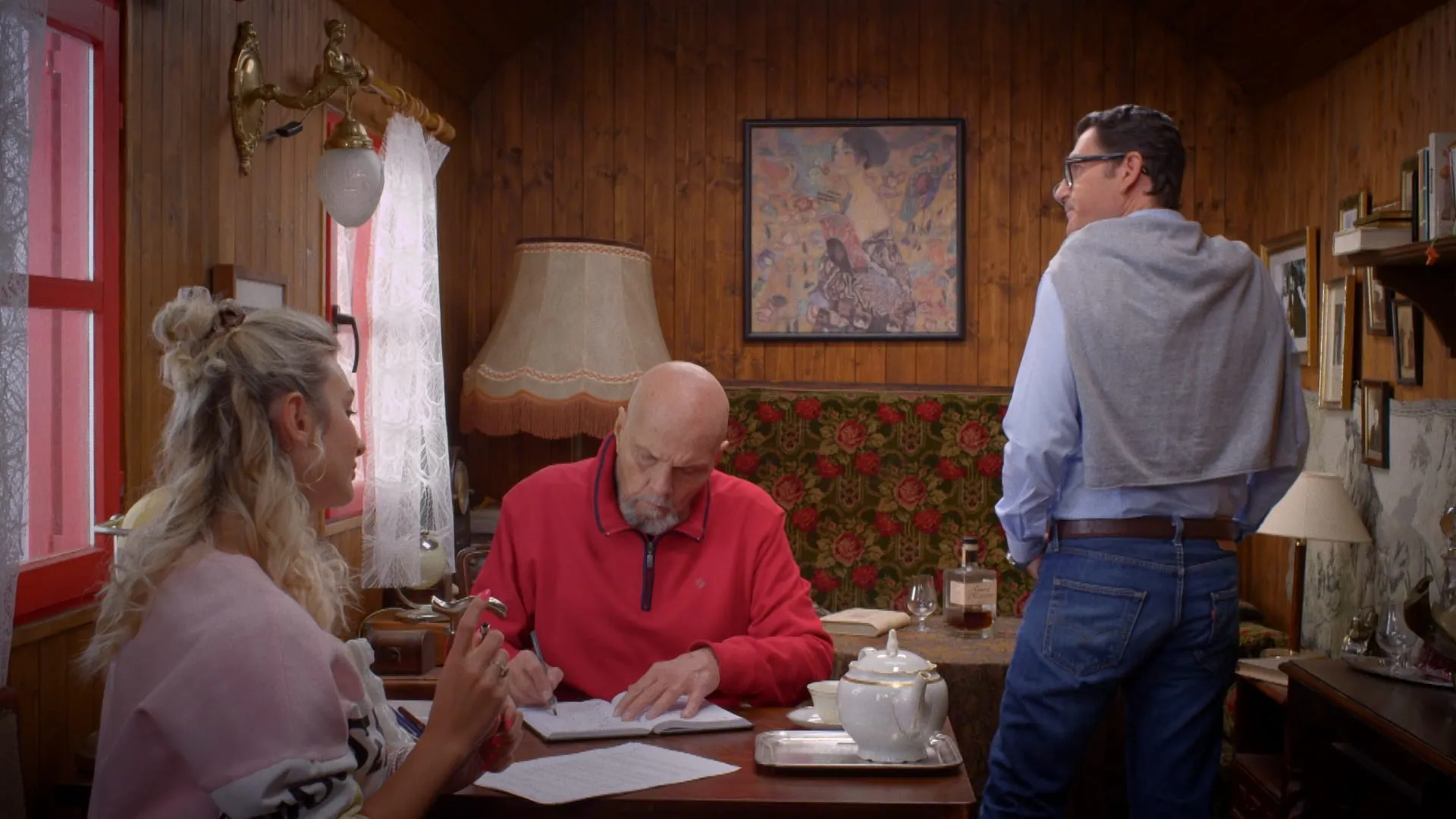In a circus caravan an older man in a red sweatshirt writes in a notebook beside a young blonde woman, and a man stands at the back