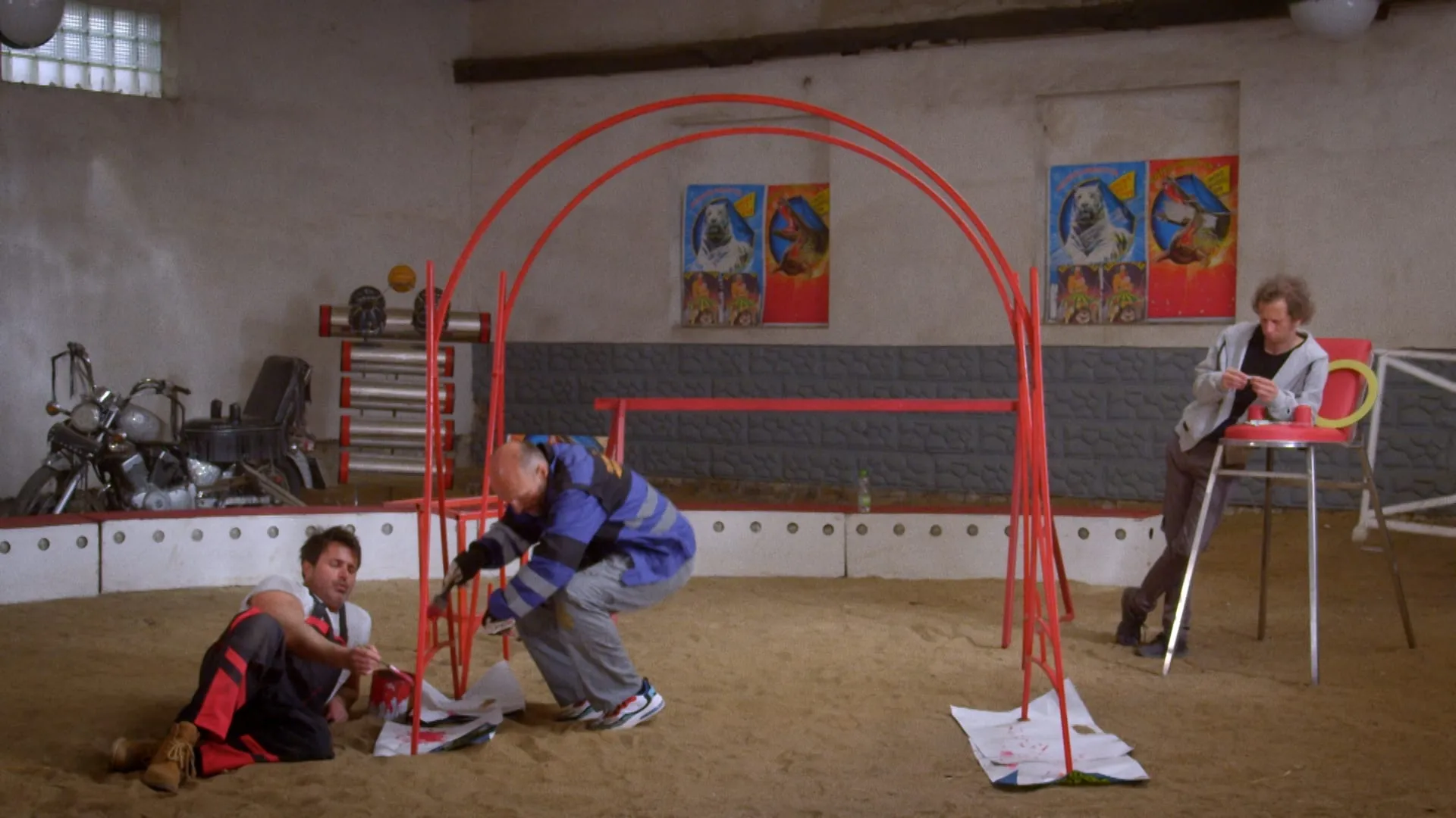Two men paint a red frame in a circus training hall while a curly-haired man sits on a bar stool