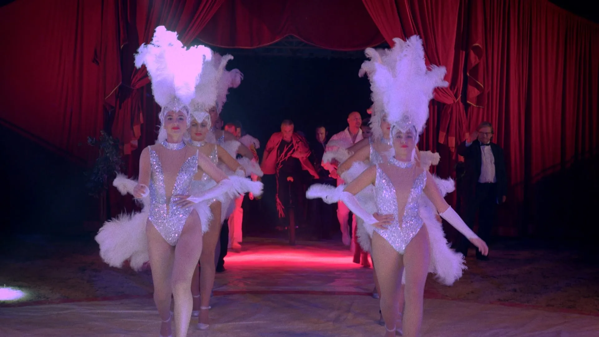 Showgirls in white feather headdresses come through the red curtain into the ring, followed by a man on a unicycle