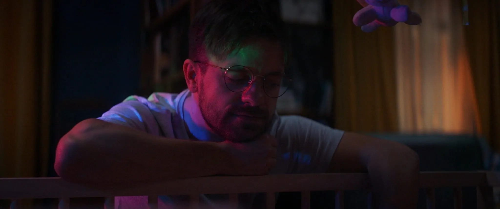 A man in glasses leans on the rail of a cot in the coloured glow of a night light and looks down at the baby