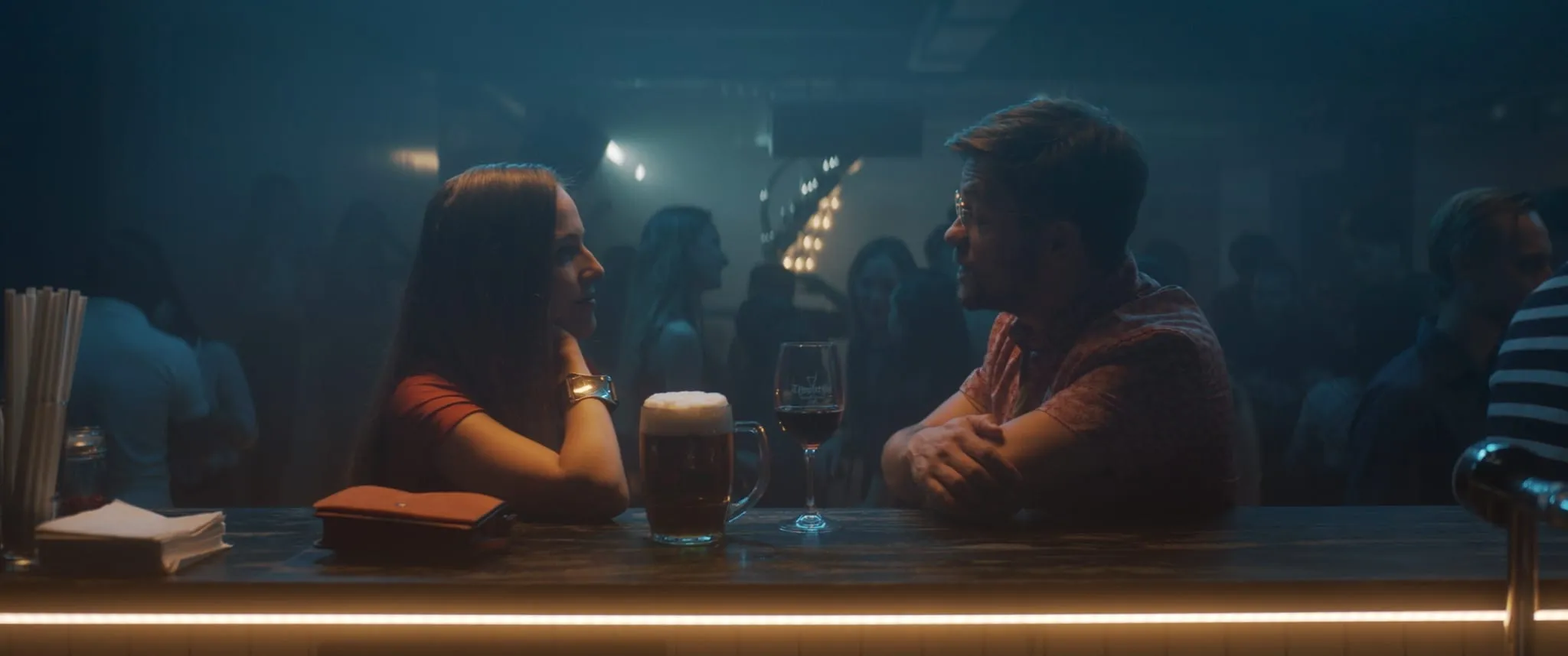 A man and a woman sit facing each other at a bar counter in a club, a beer and a glass of wine between them