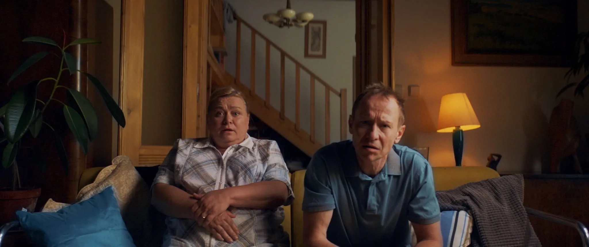 An older couple sit side by side on a living room sofa, staring ahead in disapproval