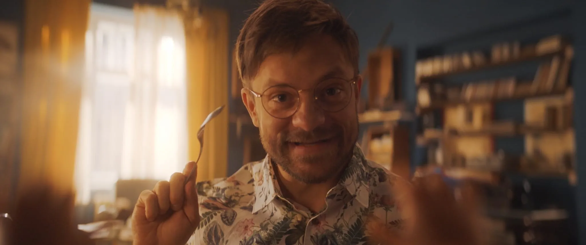 Close-up of a smiling man in glasses and a floral shirt holding a bent spoon up in front of his face