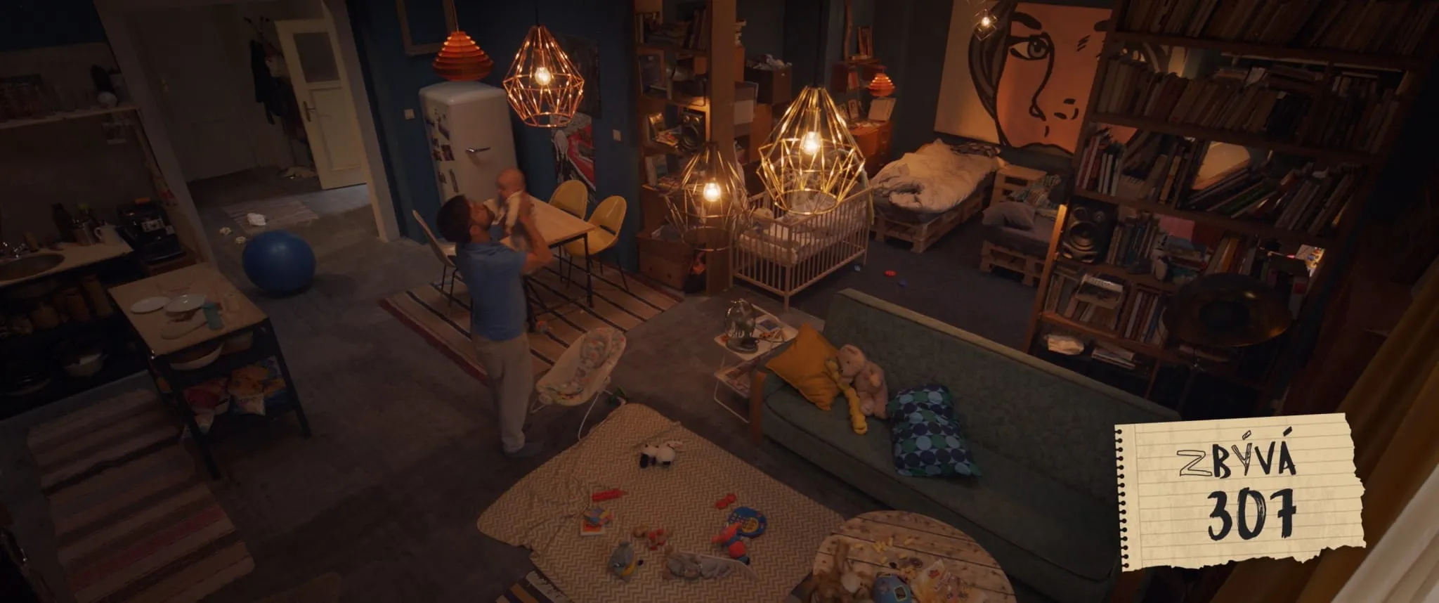 An overhead view of a cluttered flat where a man lifts a baby above his head between a cot and toys scattered on the floor
