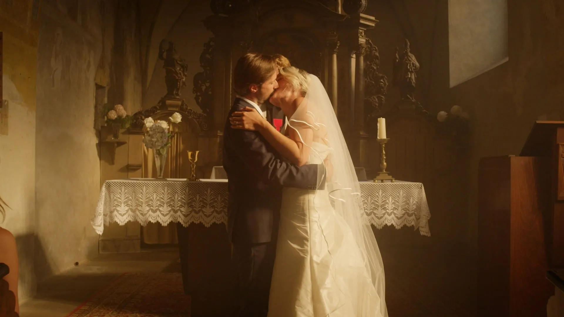 A groom and a bride in a veil kiss in front of the altar in a dim old church, a golden chalice on the altar and a candle beside it