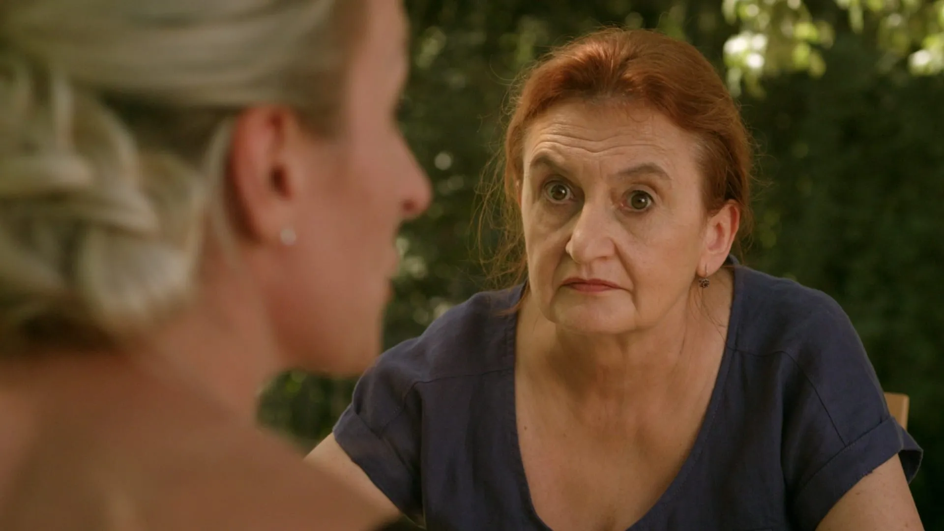 A middle-aged red-haired woman in a blue linen dress listens gravely to a blonde woman seen from behind
