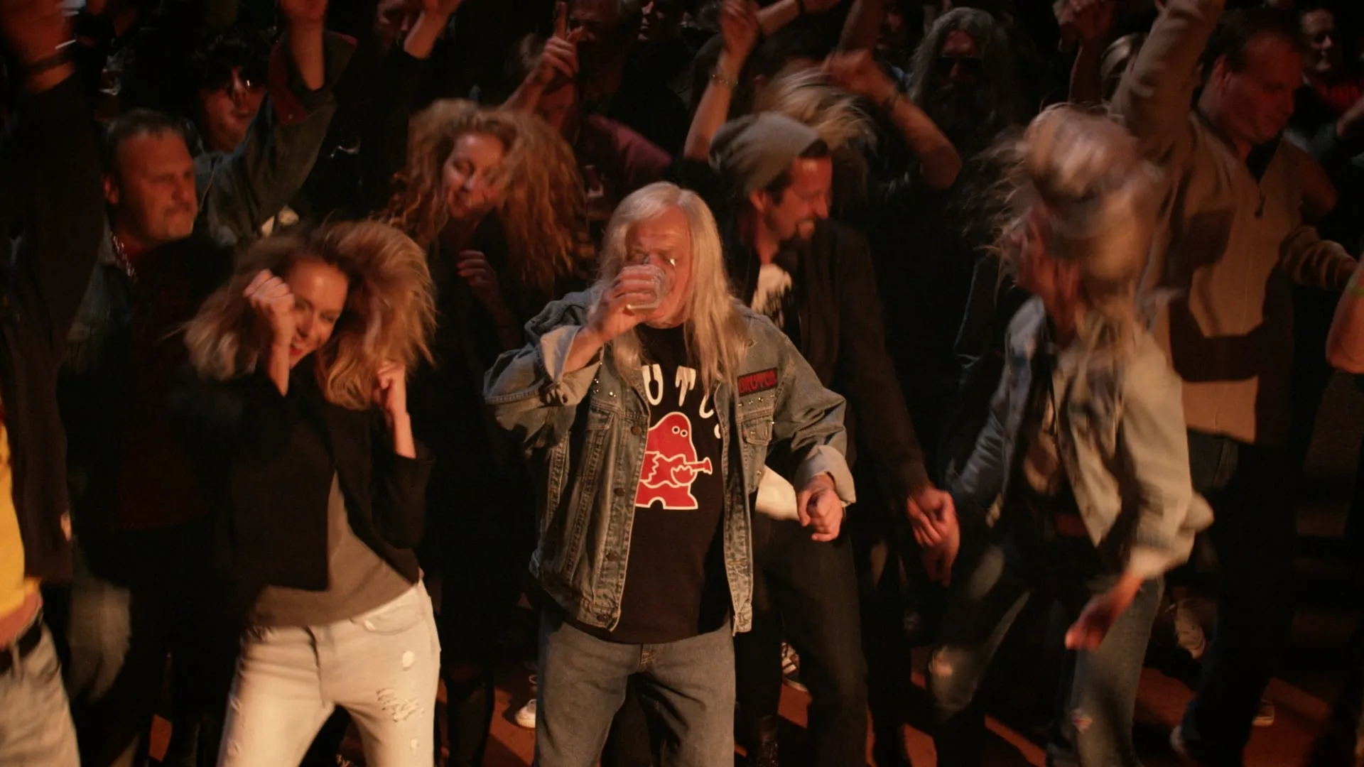 A long-haired man in a denim jacket dances on a packed dance floor, sipping from a glass as other people whirl around him