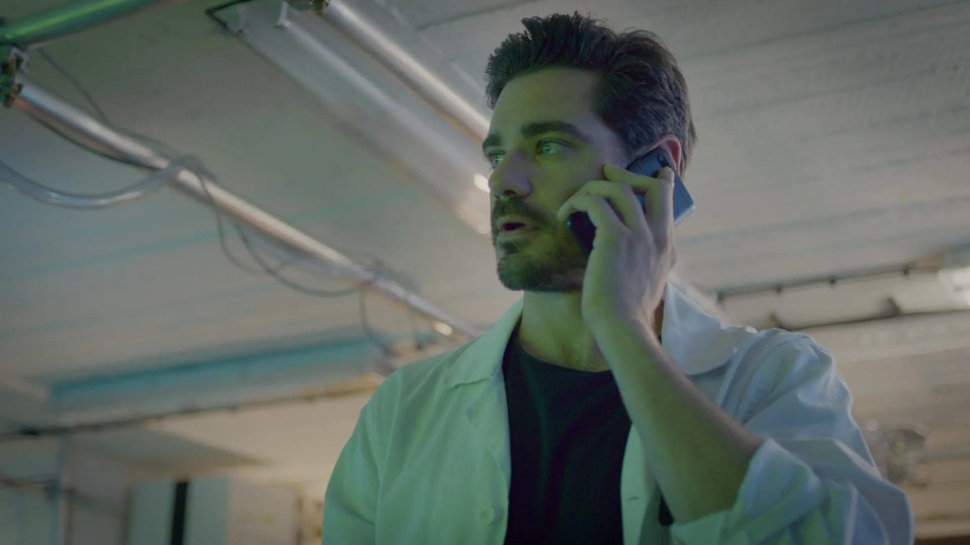 A man in a white lab coat takes a phone call under fluorescent lights, looking intently to one side