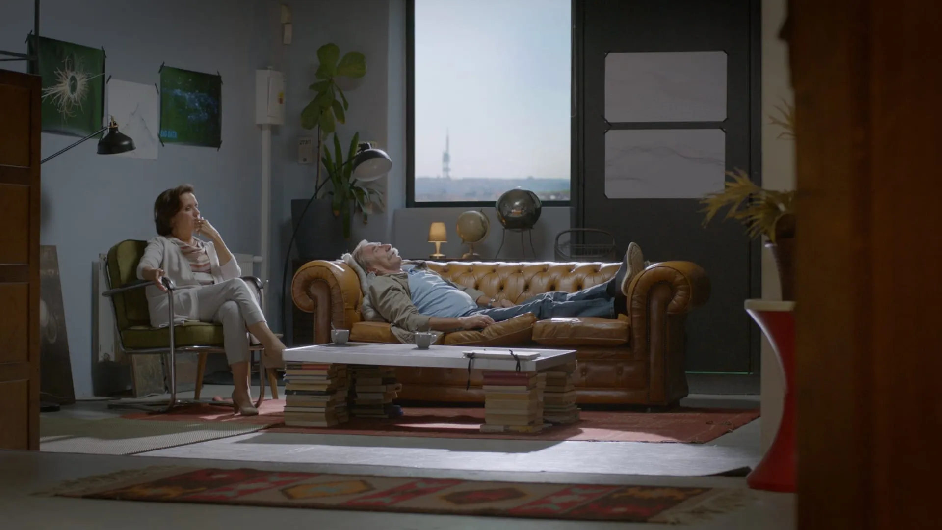 A grey-haired man lies on a leather sofa staring at the ceiling while a woman in a white suit watches him from an armchair