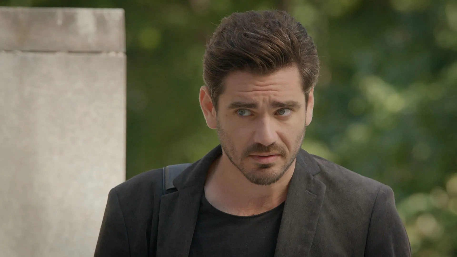 Close-up of a young man in a blazer over a black T-shirt squinting suspiciously to one side outdoors by a stone wall