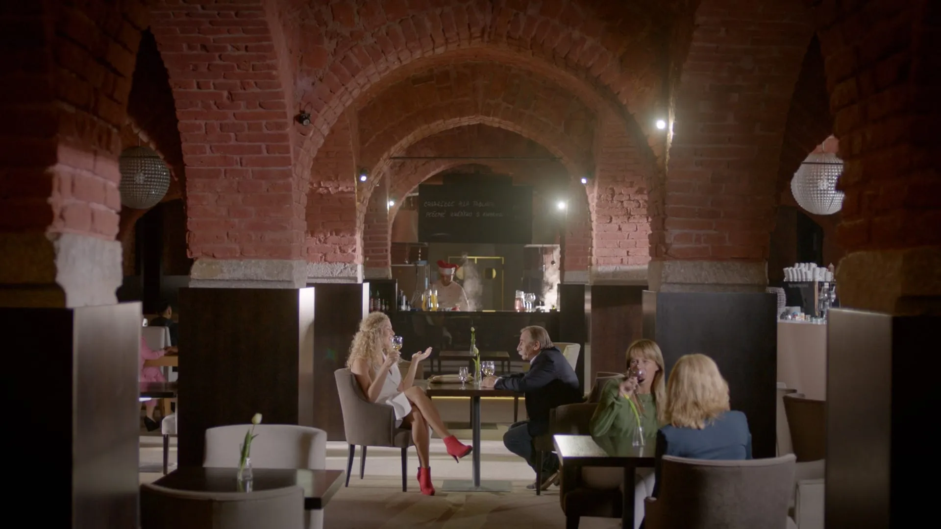 In a cellar restaurant with brick vaults a curly-haired blonde woman with a glass of wine talks to a man at the next table