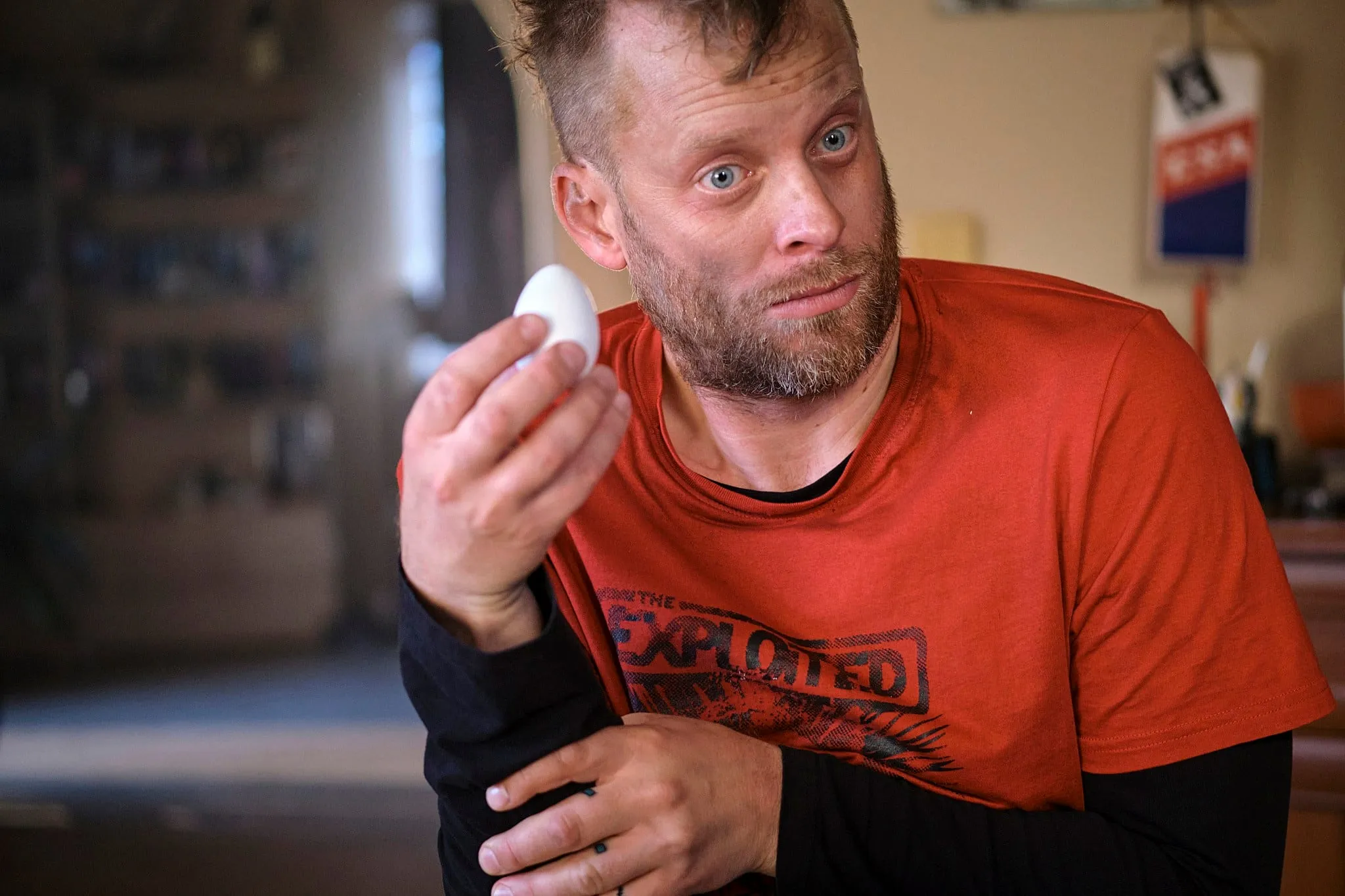 A dishevelled bearded man in a red T-shirt holds out a white egg and stares into the camera in bewilderment