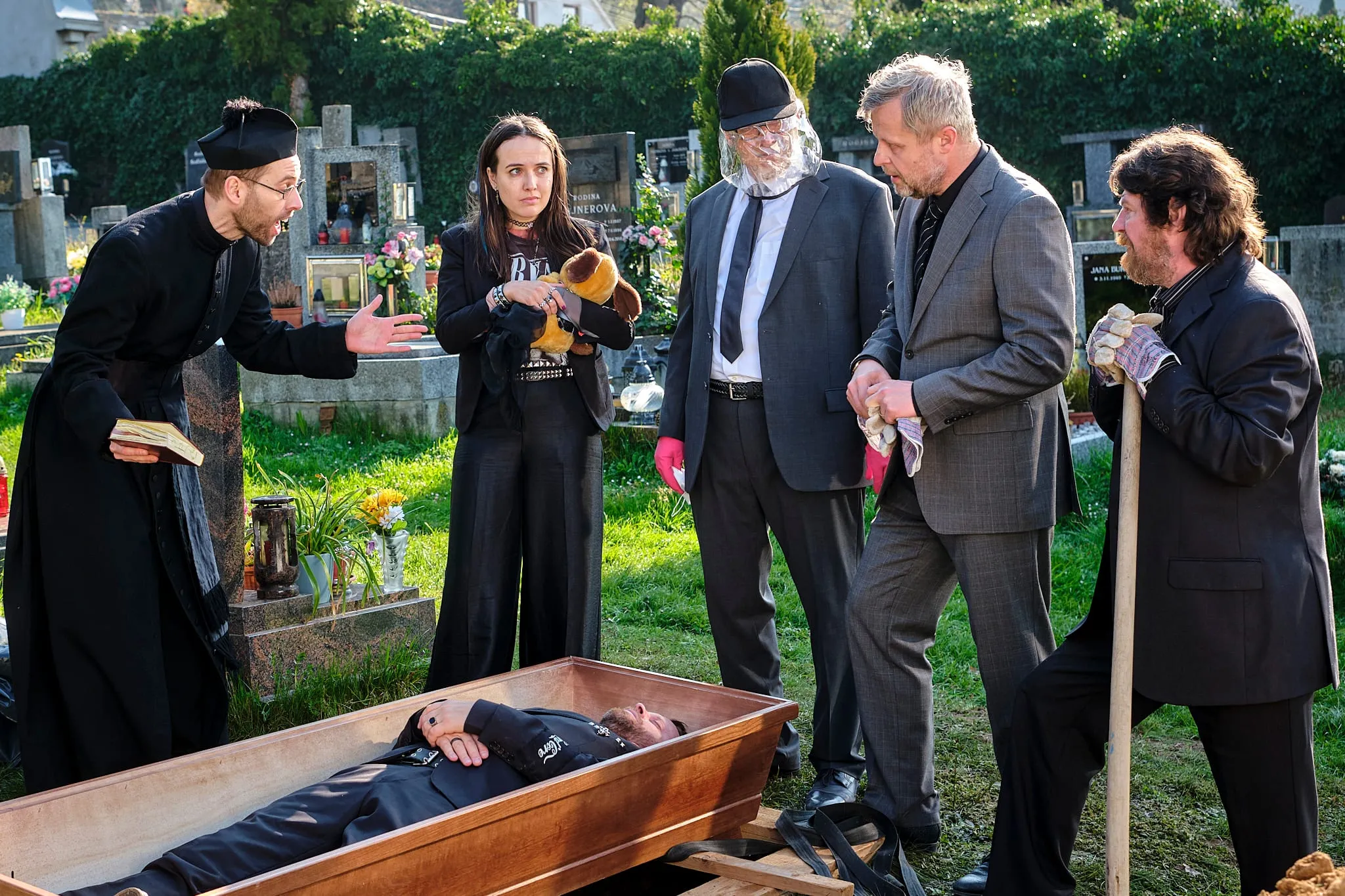 At a cemetery a priest, a young woman with a toy dog and men in suits stand around an open coffin with a man in it