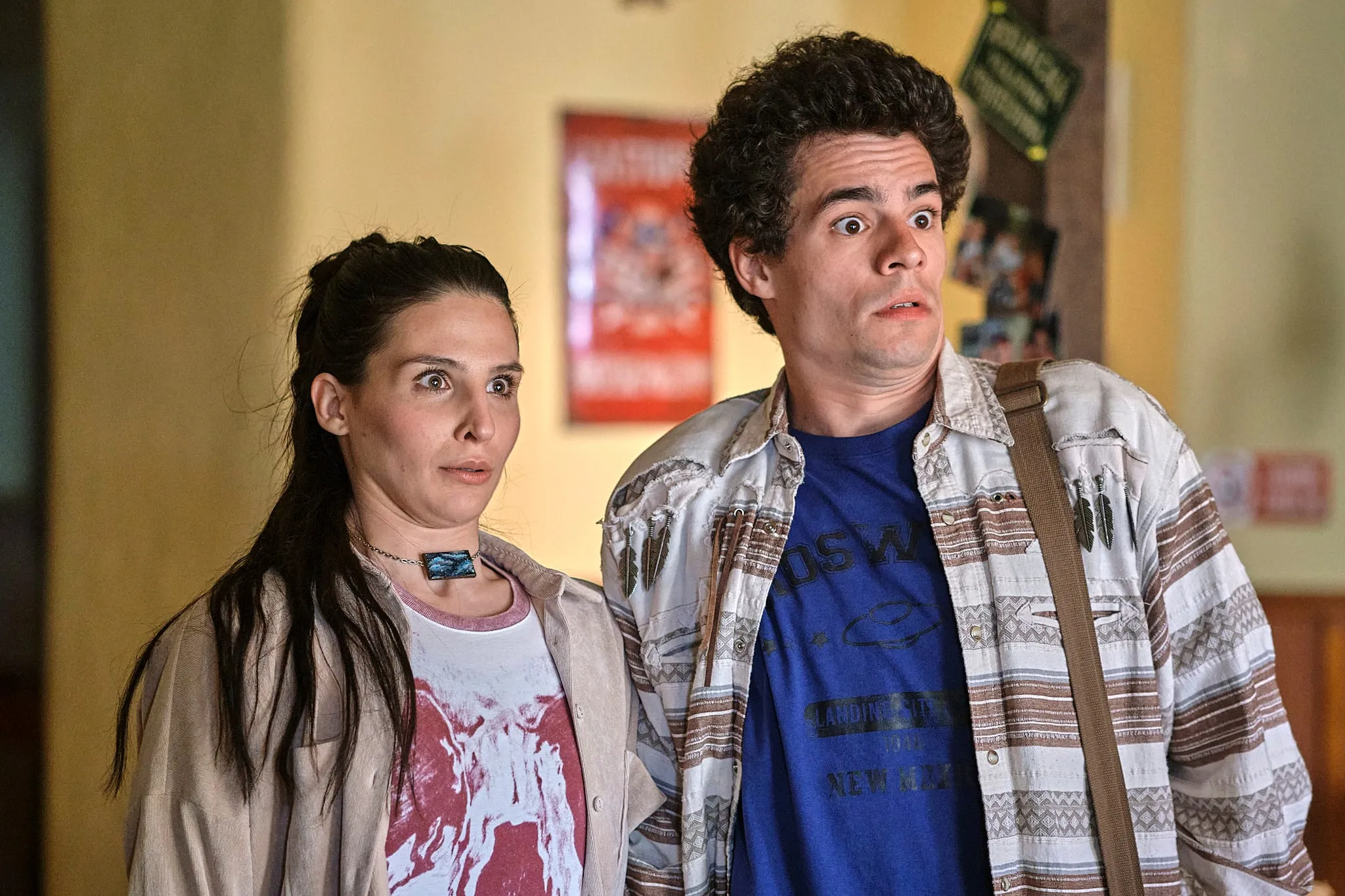 A dark-haired woman and a curly-haired young man stand side by side indoors, staring ahead in astonishment