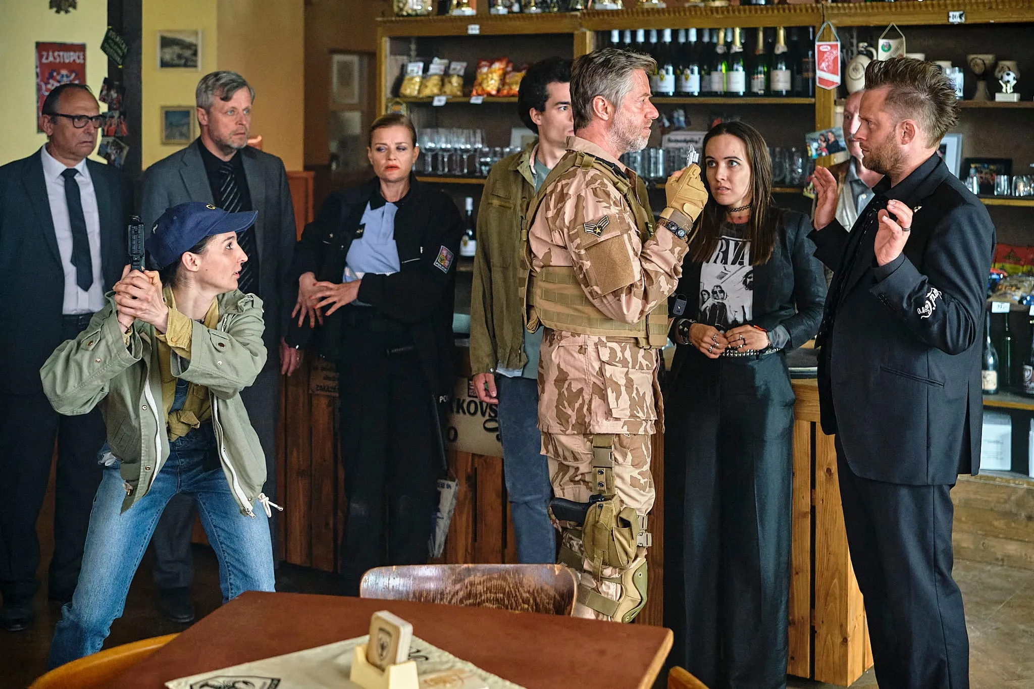 In a pub a woman in a cap aims a pistol at a man in desert camouflage while a policewoman and other villagers watch