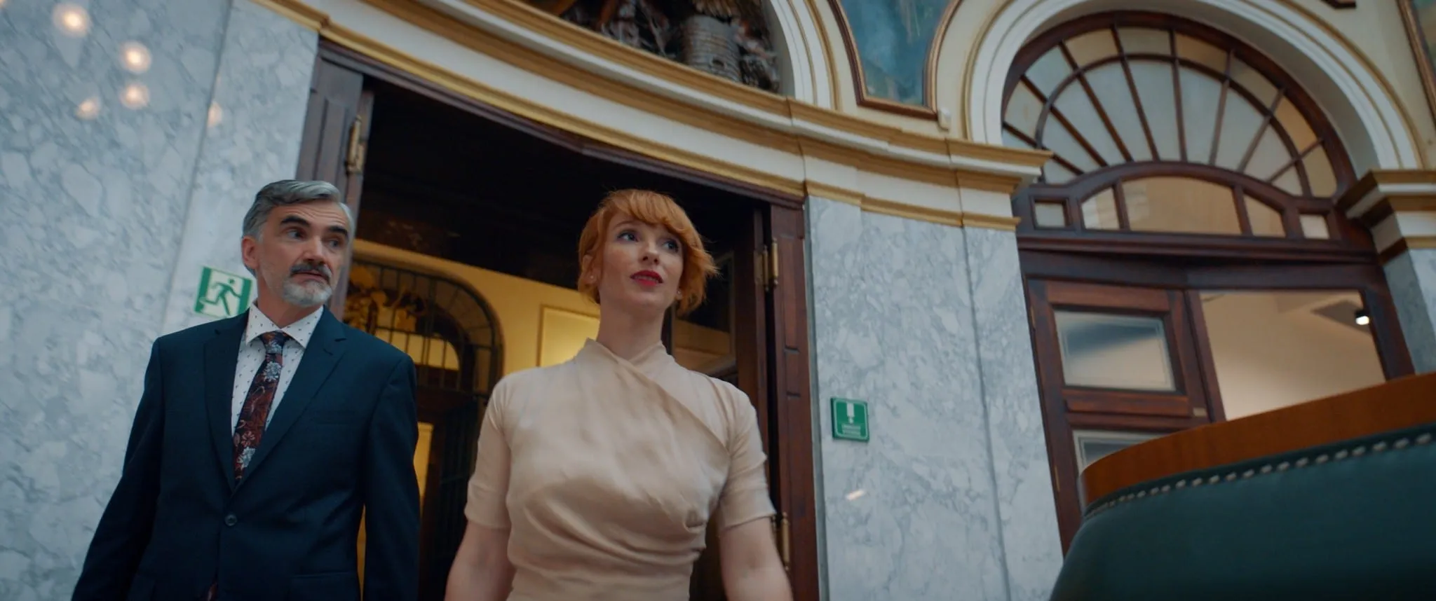 A grey-haired man in a suit and a red-haired woman in a cream dress step out of a doorway in a marble hall, both looking up