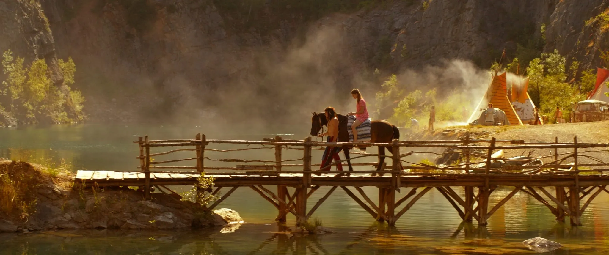 A man leads a horse carrying a girl in a pink dress across a wooden bridge over a lake, tipis on the bank