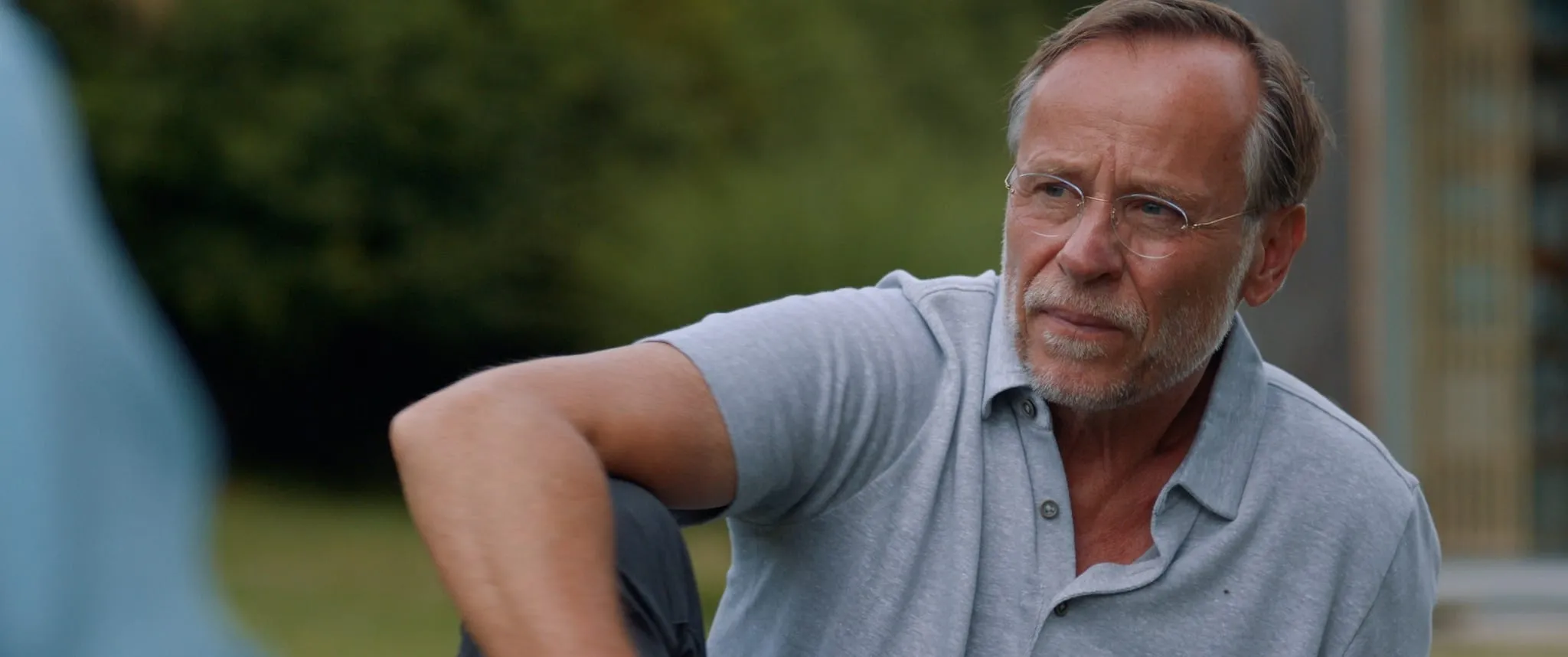 An older man in glasses and a grey polo shirt sits in a garden, looking thoughtfully to one side