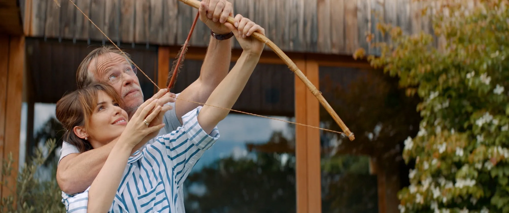 A woman hugs a man from behind as he draws a homemade bow and arrow, both looking upwards