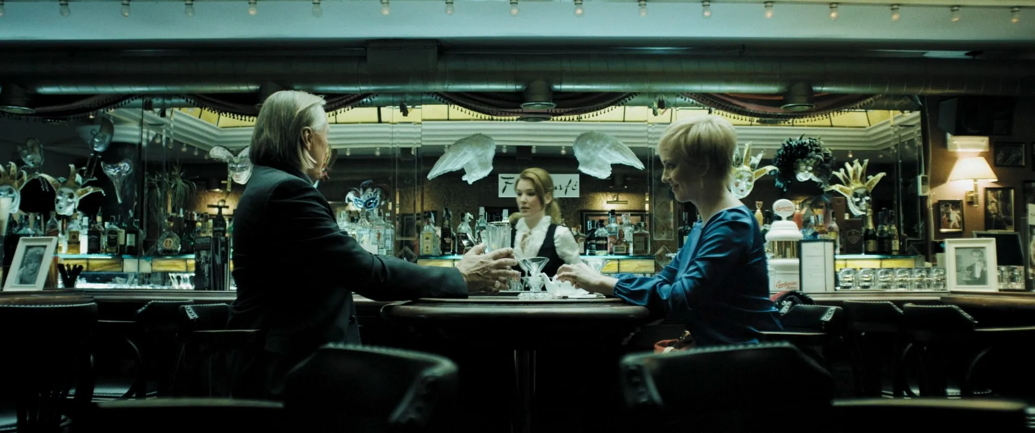 A grey-haired man in a suit sits with his back to the camera at a bar opposite a blonde woman in a blue dress, Venetian masks on the walls