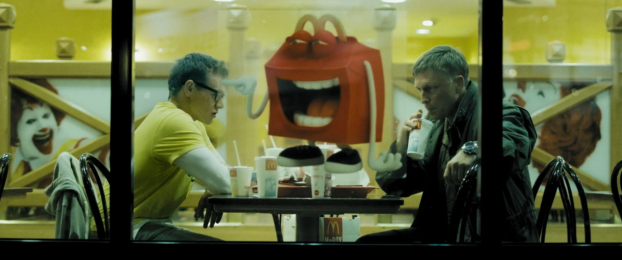 A view through a window into a fast-food restaurant where two men sit facing each other with drinks and paper bags