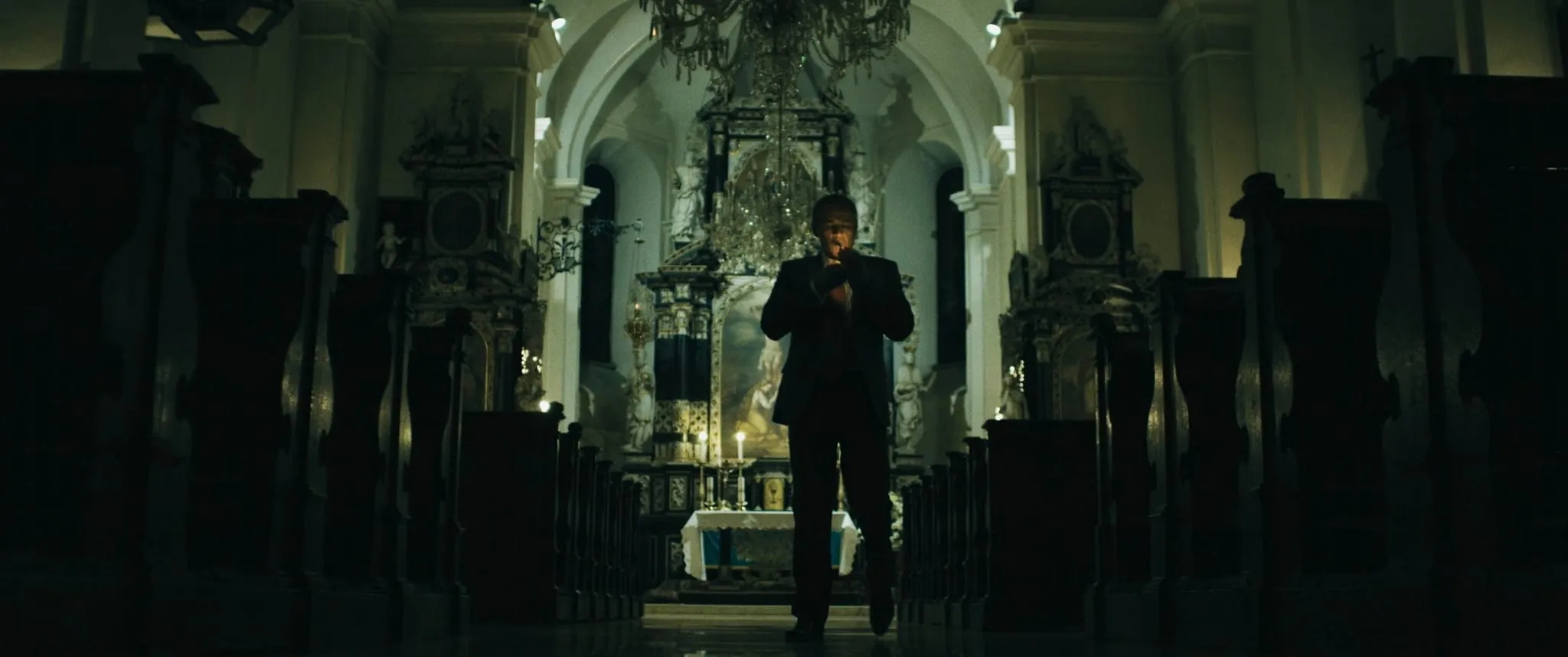 A man in a dark suit lights a cigarette in the aisle of a dimly lit baroque church