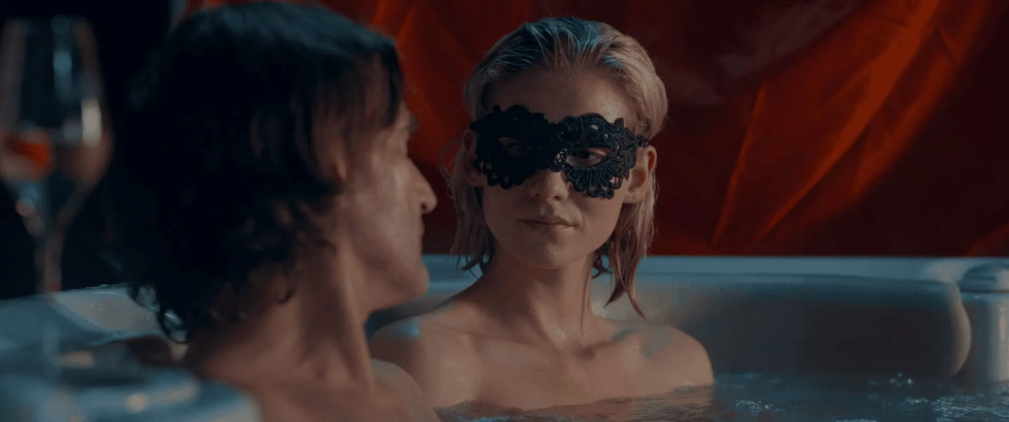 A woman in a black lace mask sits in a hot tub facing a man, red fabric glowing behind them
