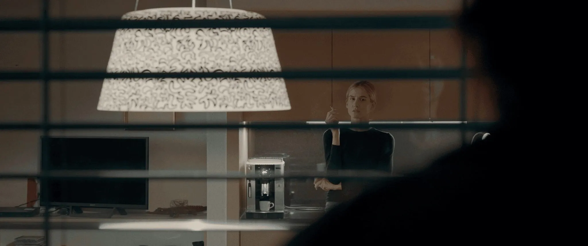 Seen through window blinds, a blonde woman stands smoking in a flat while a dark figure watches
