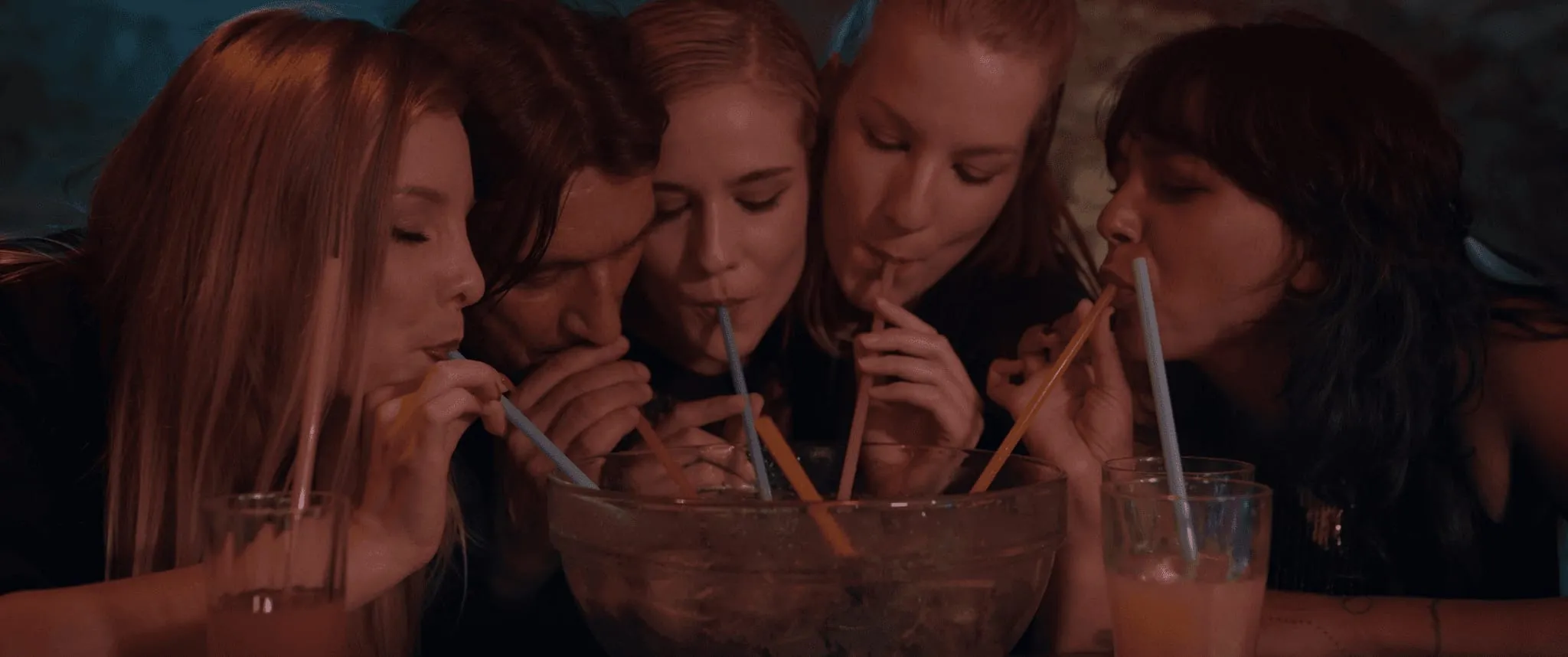 Five young women lean over a large glass bowl and drink through straws in a dimly lit bar