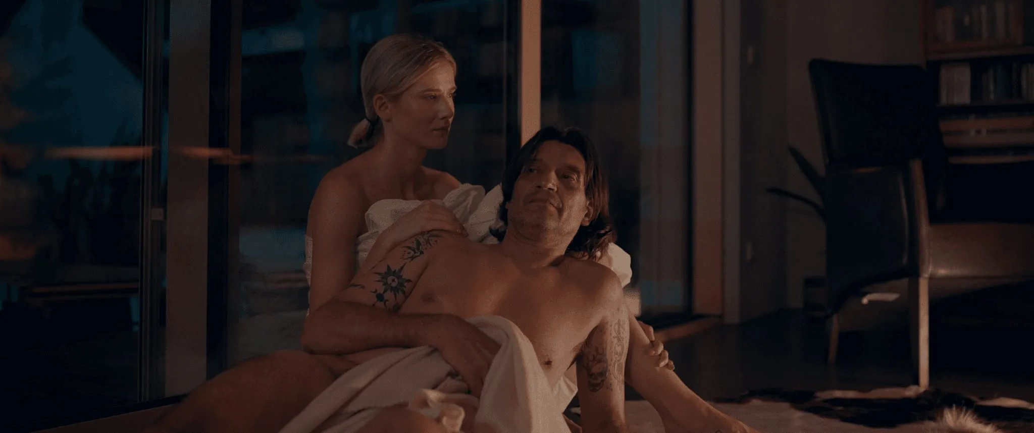 A blonde woman wrapped in a sheet holds a tattooed man from behind as they sit on the floor by a glass wall