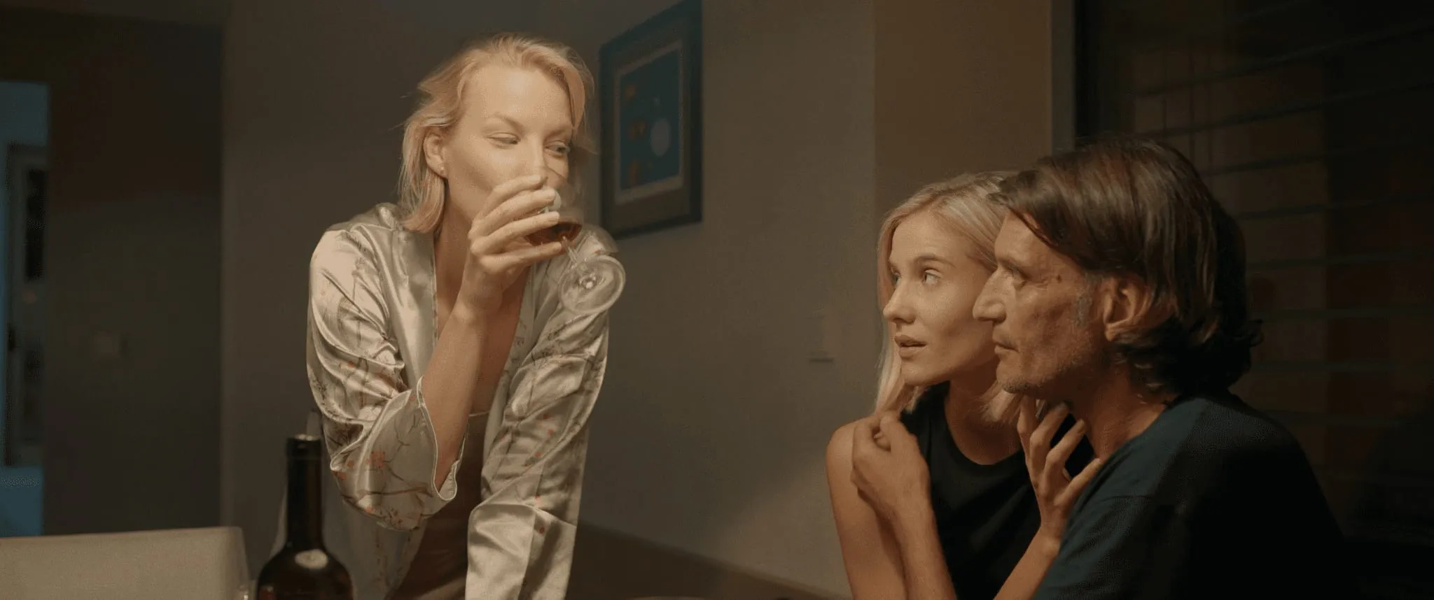 A woman in a satin robe drinks wine at a table while, on the right, a younger blonde woman and a long-haired man sit close together