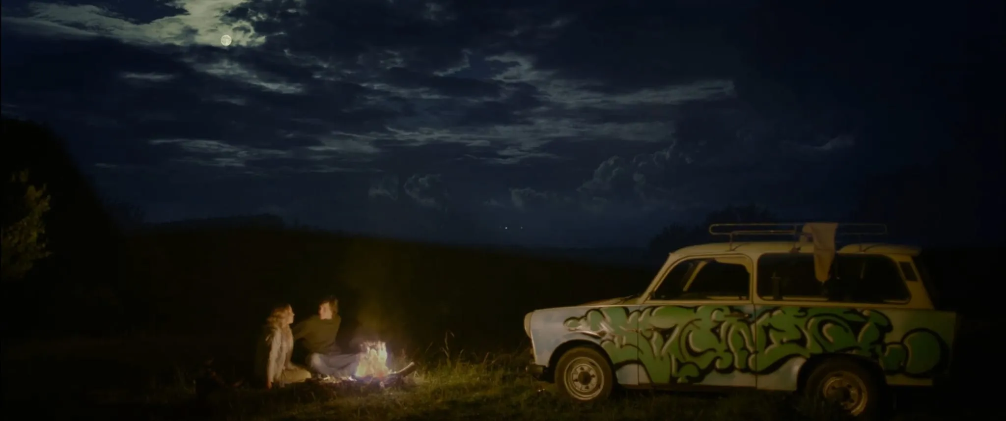 Mladý pár sedí v noci u ohně na louce, vedle stojí trabant kombi pomalovaný zeleným graffiti