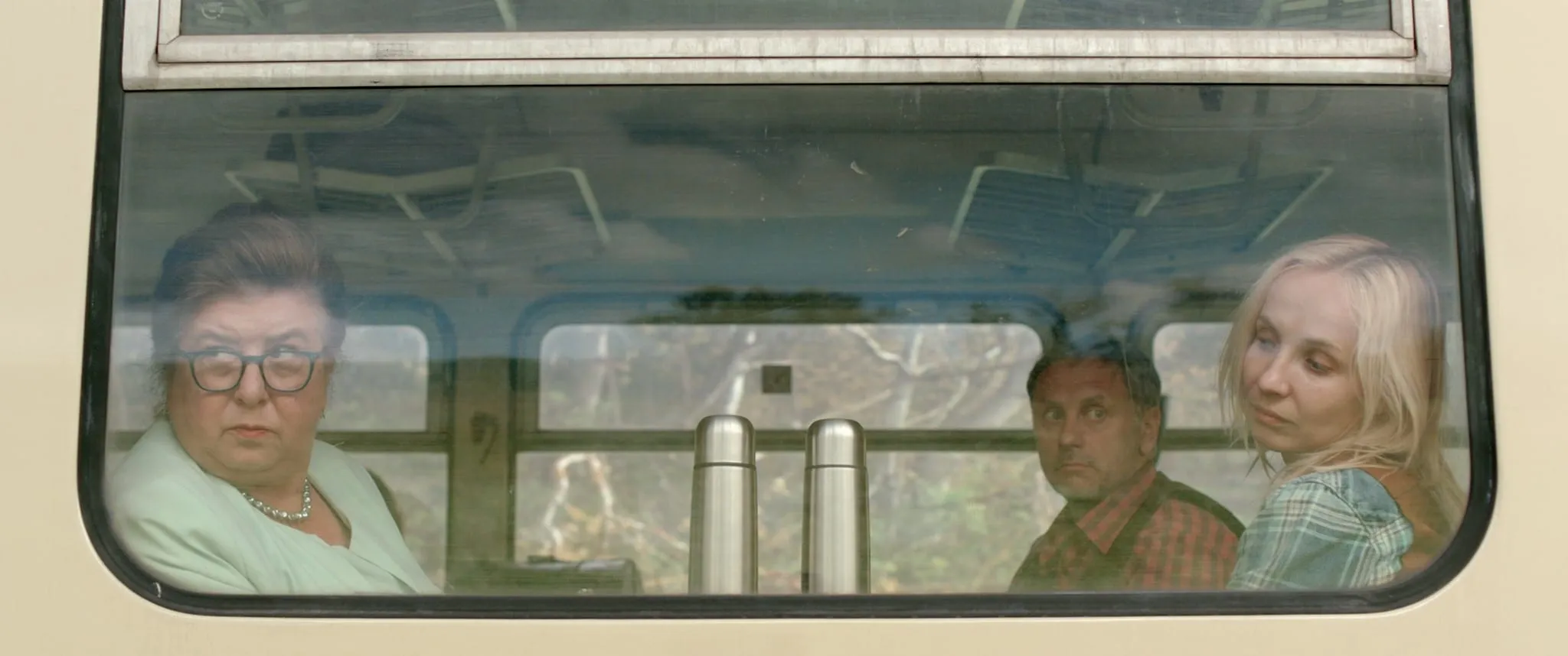 An older woman in glasses, a man in a checked shirt and a young blonde woman seen through a railcar window