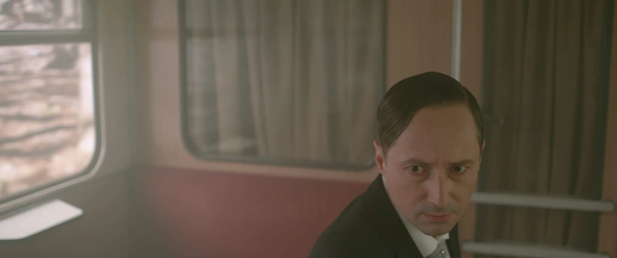 A man with slicked-back hair in a dark suit sits in a carriage and looks up with a frown