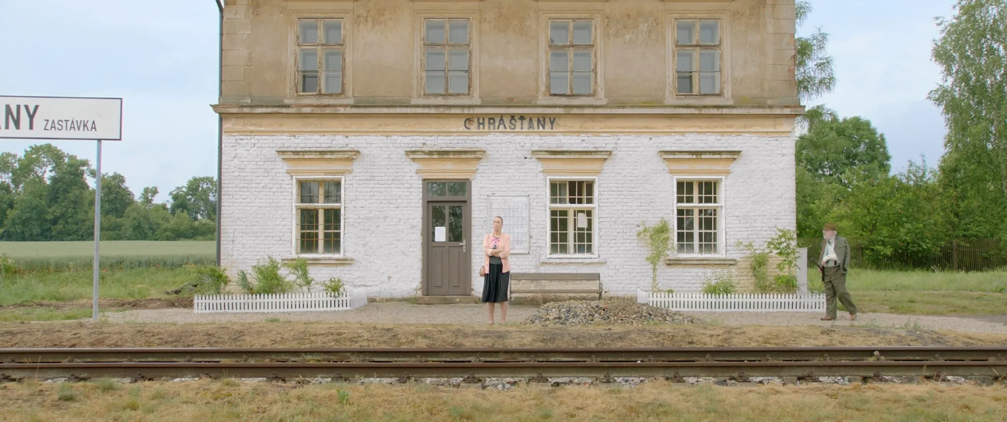 A woman stands in front of the small country station at Chrášťany while an older man in a hat walks up on the right