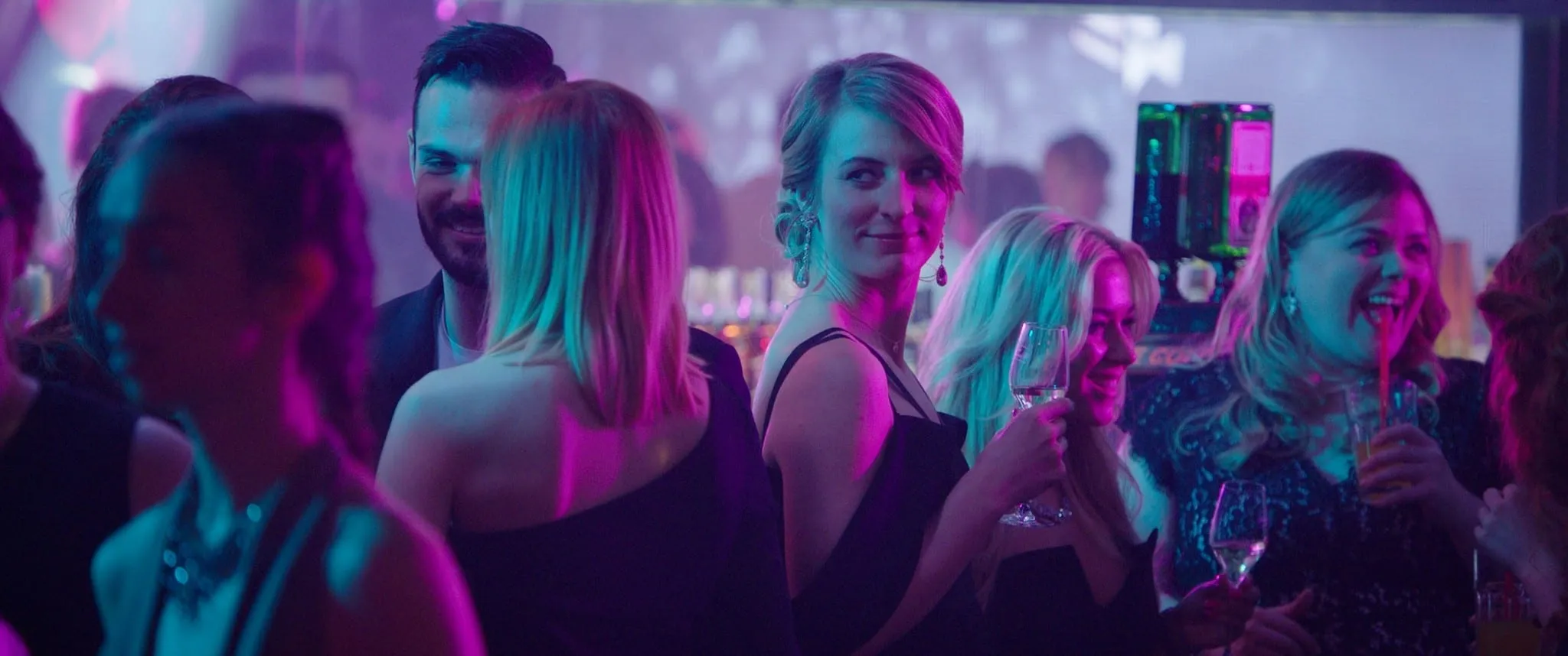 A short-haired blonde woman in a black dress turns towards the camera with a glass of wine in a colour-lit club