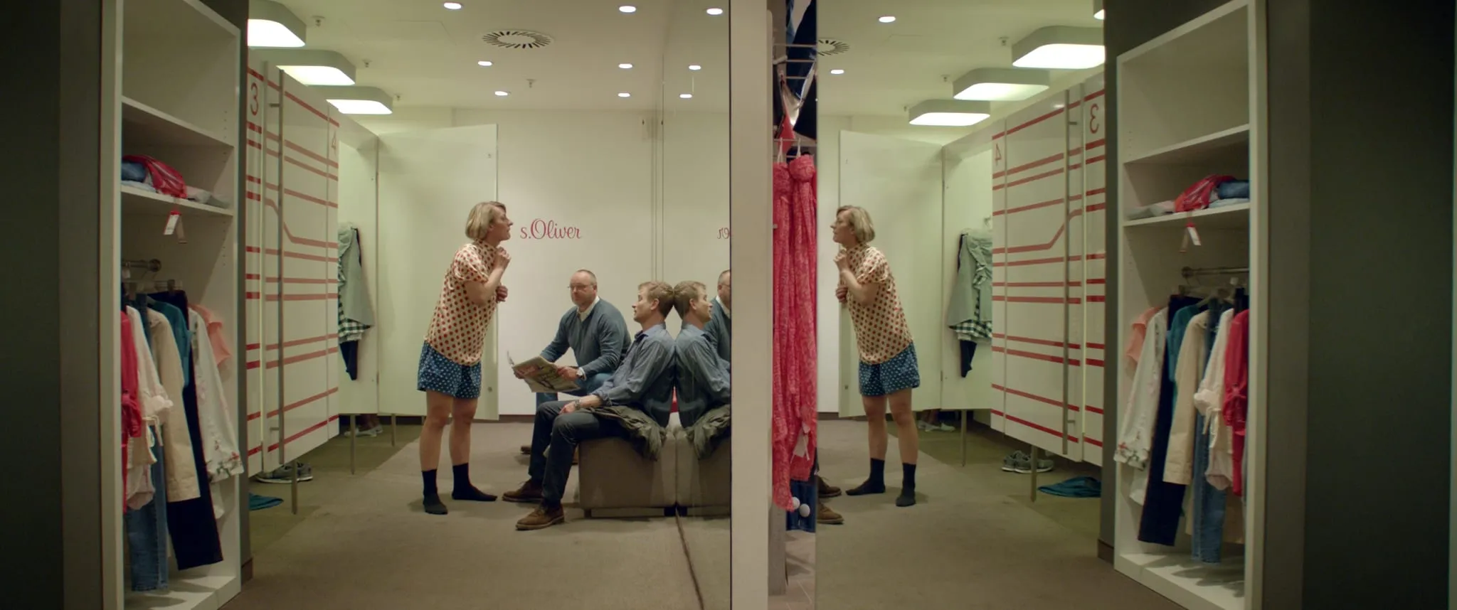 A short-haired blonde woman stands in a shirt and boxer shorts before a fitting room mirror, two men sitting on a bench behind