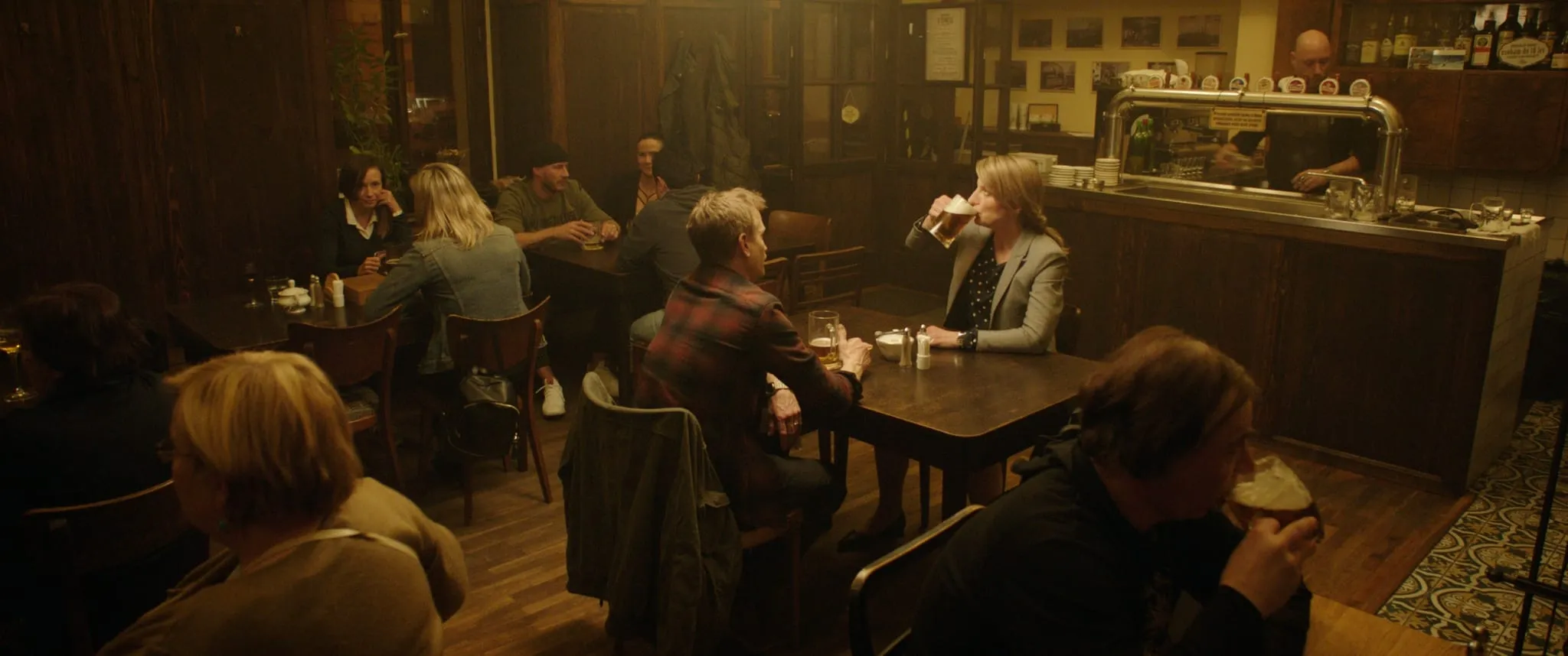 In a wood-panelled pub a blonde woman in a grey blazer drinks beer at a table opposite a man in a checked shirt