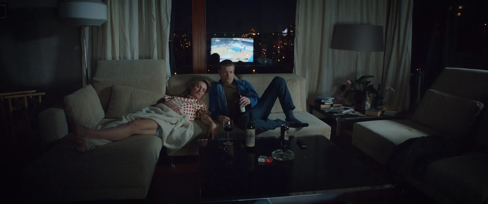 A woman in a polka-dot shirt sleeps on a sofa beside a man holding a beer bottle, basketball on the television behind