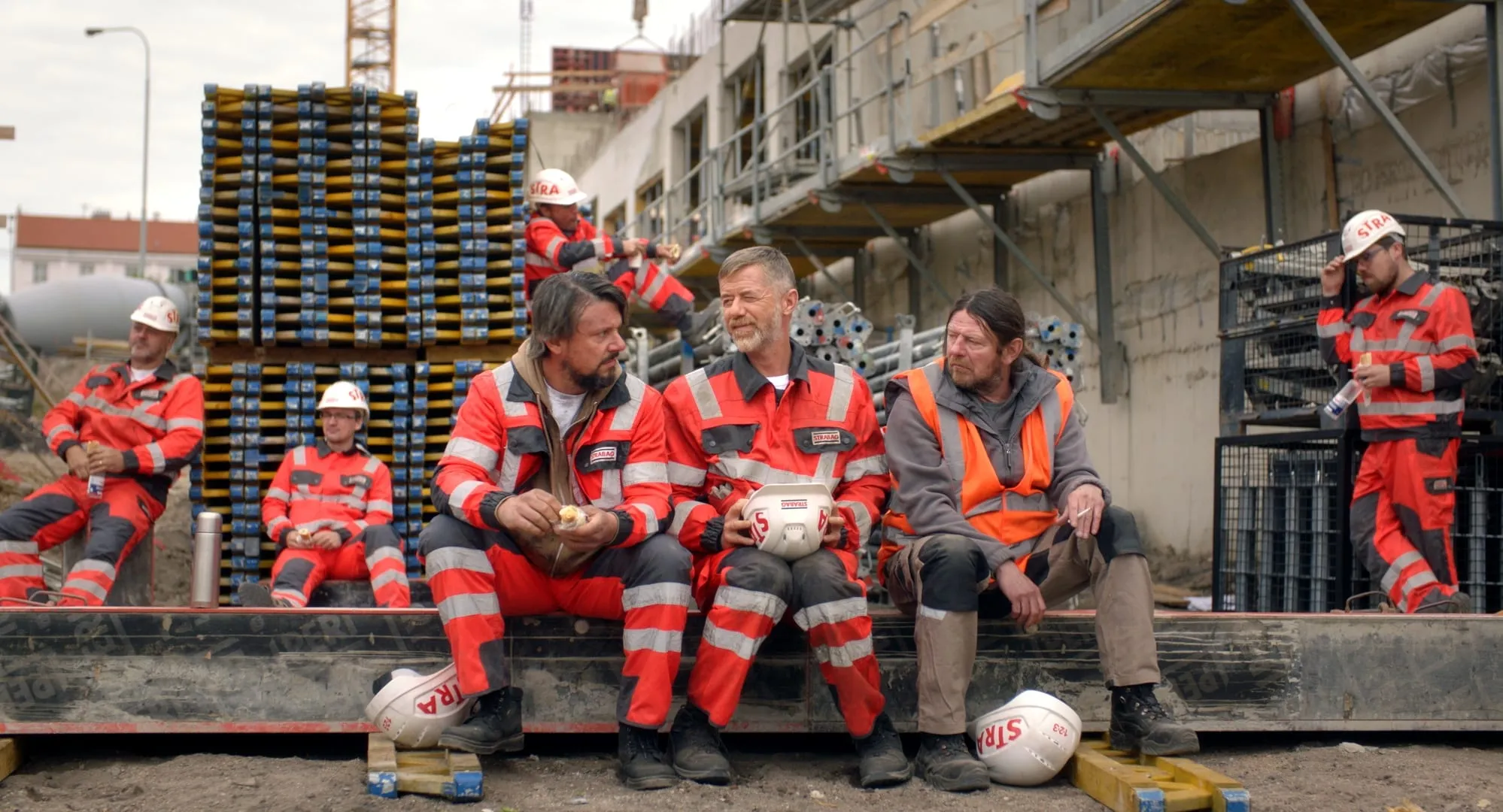 Workers in red high-visibility overalls sit on a beam during a break at a building site, hard hats on the ground