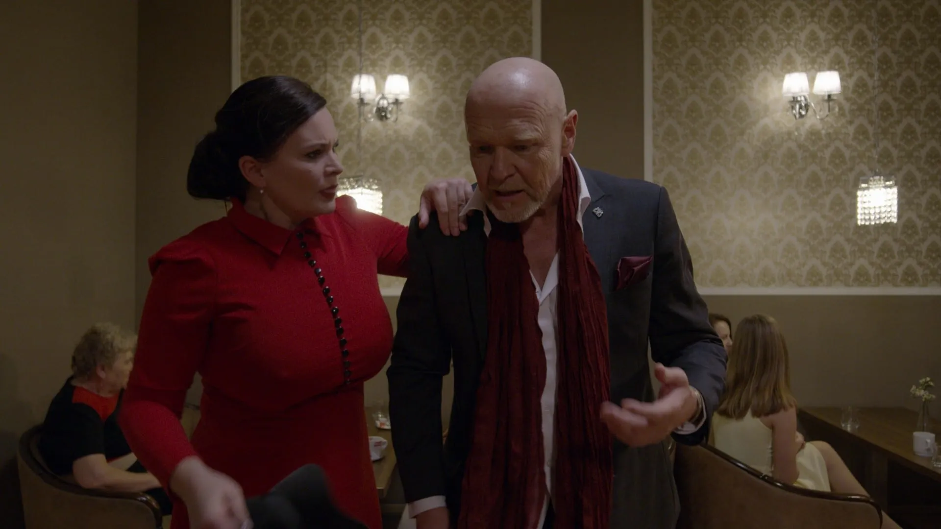 A woman in a red dress grips a man in a scarf by the shoulder and talks at him angrily in a hotel café