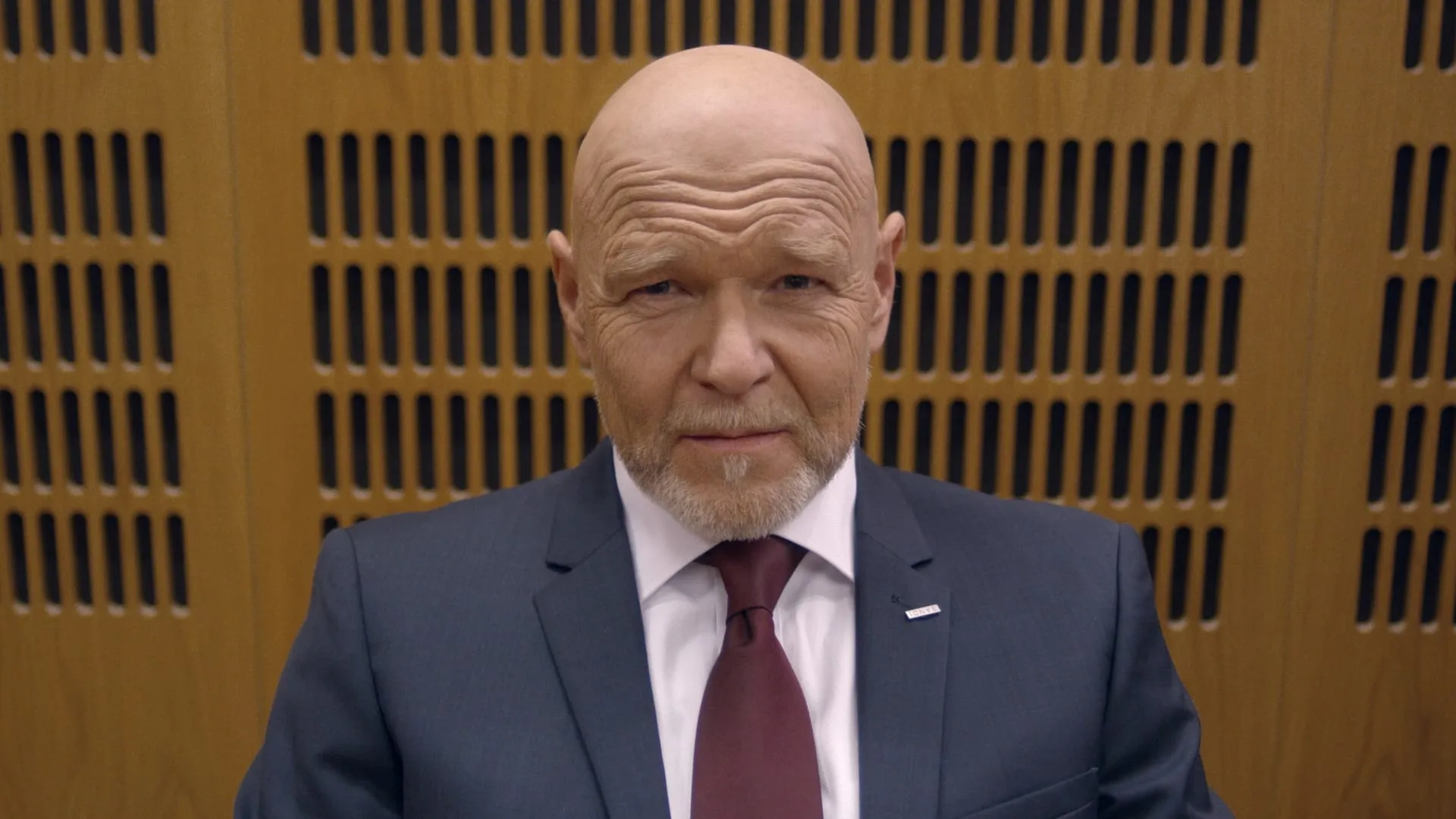 Portrait of a bald bearded man in a blue suit and burgundy tie in front of a wood-panelled wall