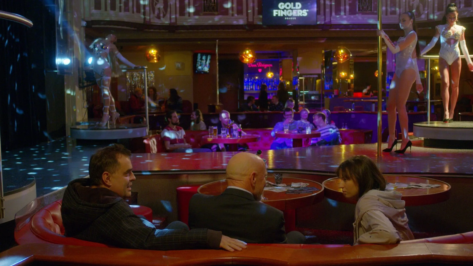 Two men and a young woman sit in a red booth in a nightclub, pole dancers on the podiums behind them