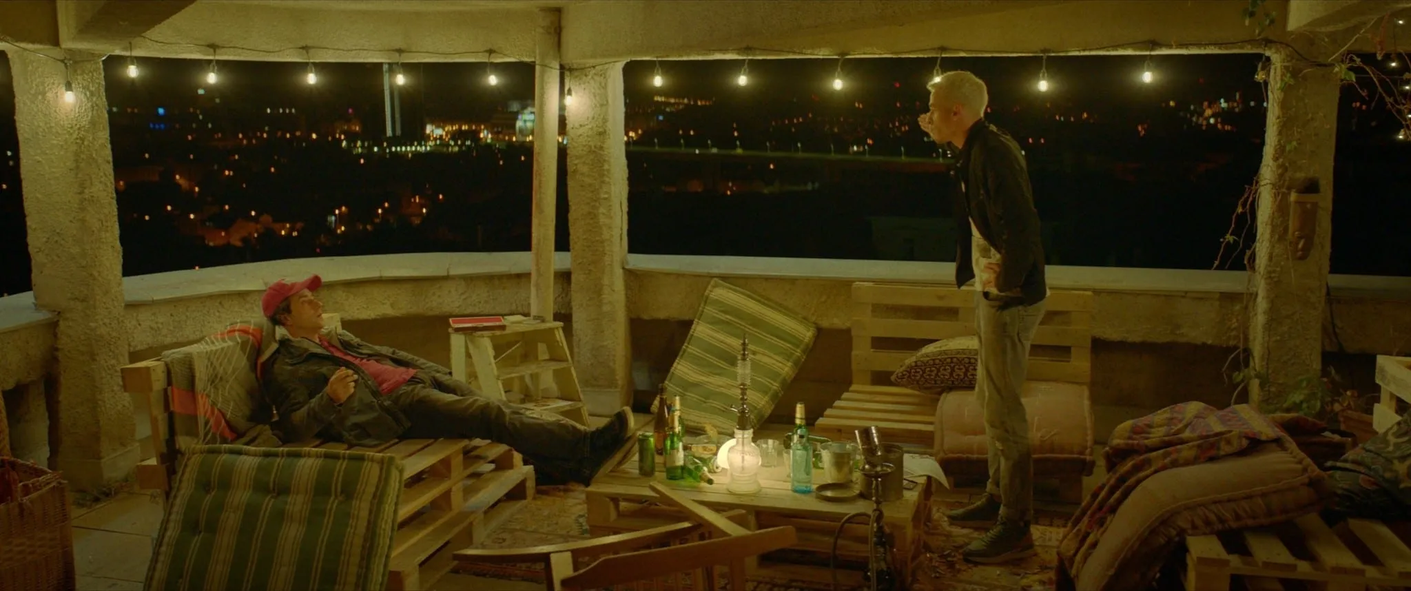 A man in a red cap lies on a pallet lounger on a terrace while a blond man stands opposite him, the city lit up at night behind them