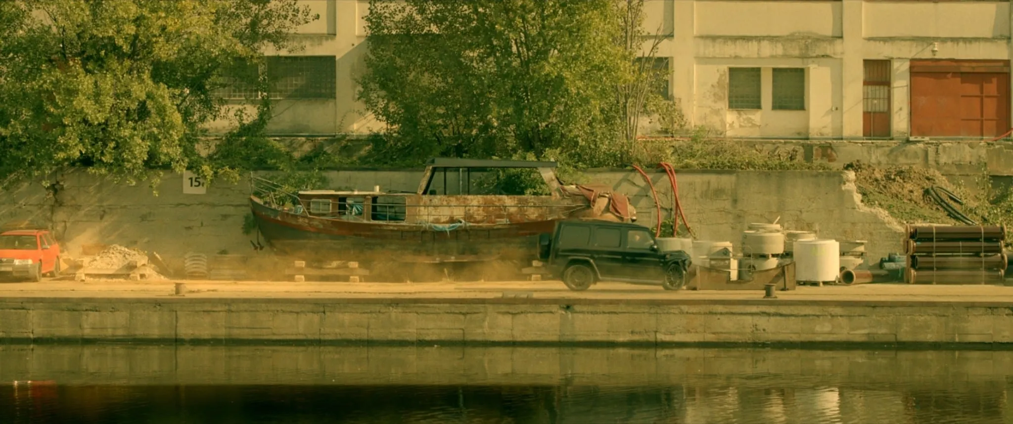 A black SUV speeds along a riverside quay past a rusty boat hauled up on the bank, raising dust behind it